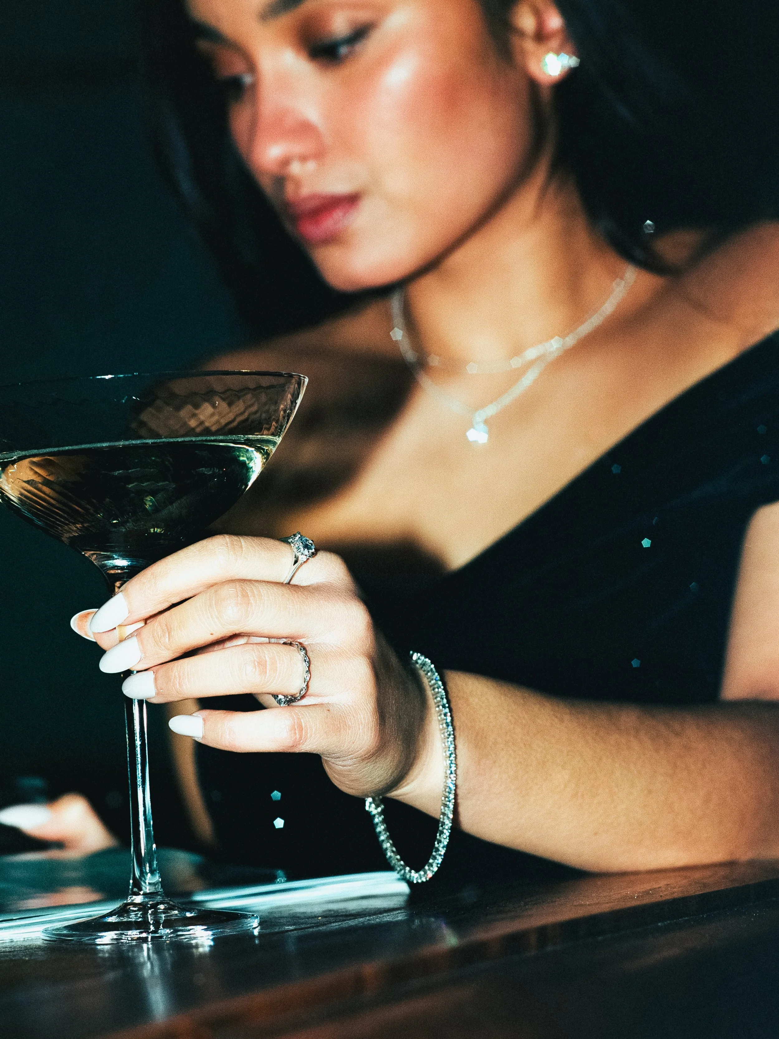 A woman with dark hair and earrings holding a glass of dark liquid, dressed in black with jewelry, in a dimly lit setting.
