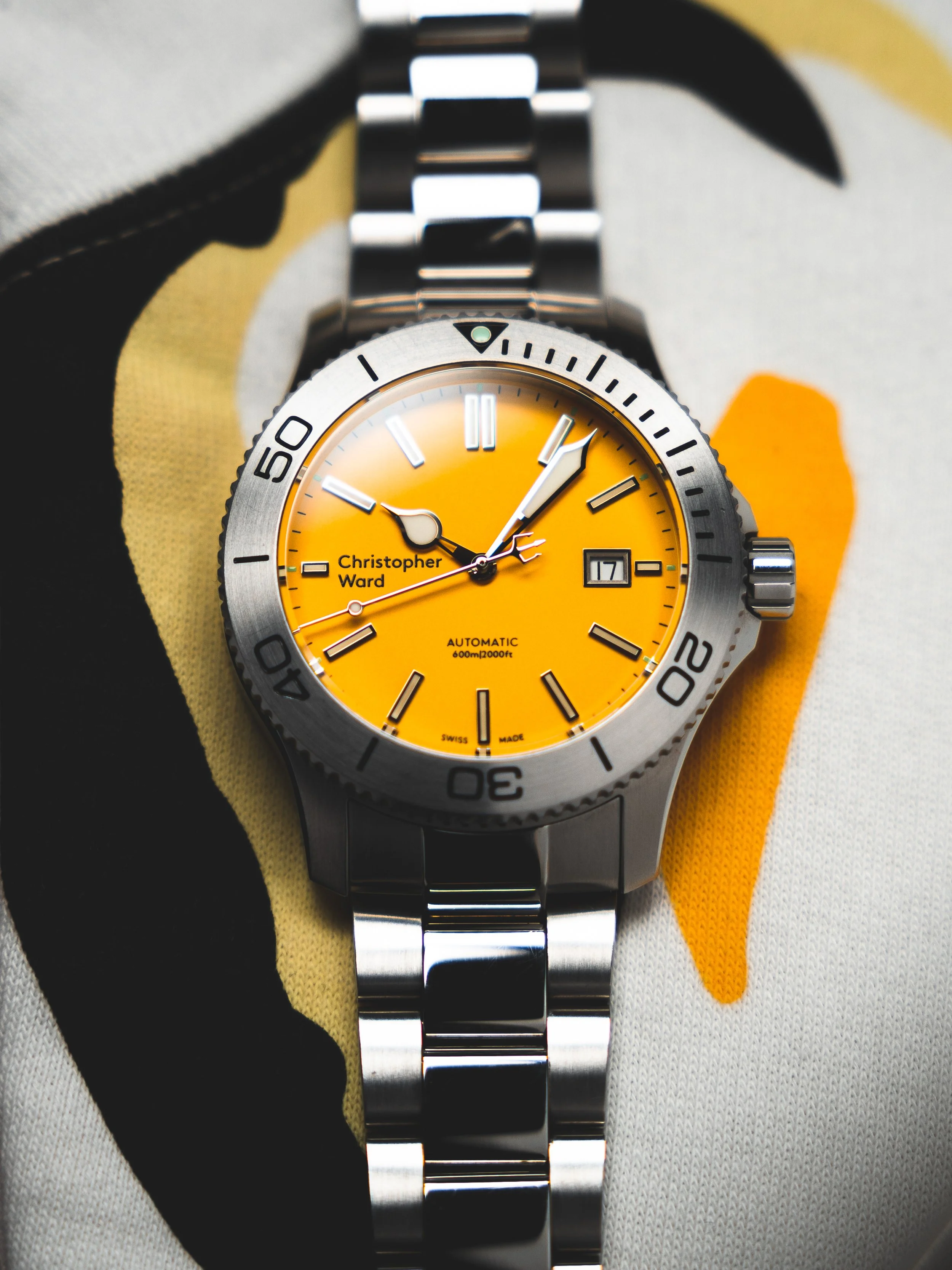 A silver wristwatch with a yellow dial, black hour markers, and a stainless steel band. The watch displays the day and date, with the time showing approximately 2:07. The brand name on the dial reads "Christopher Ward".