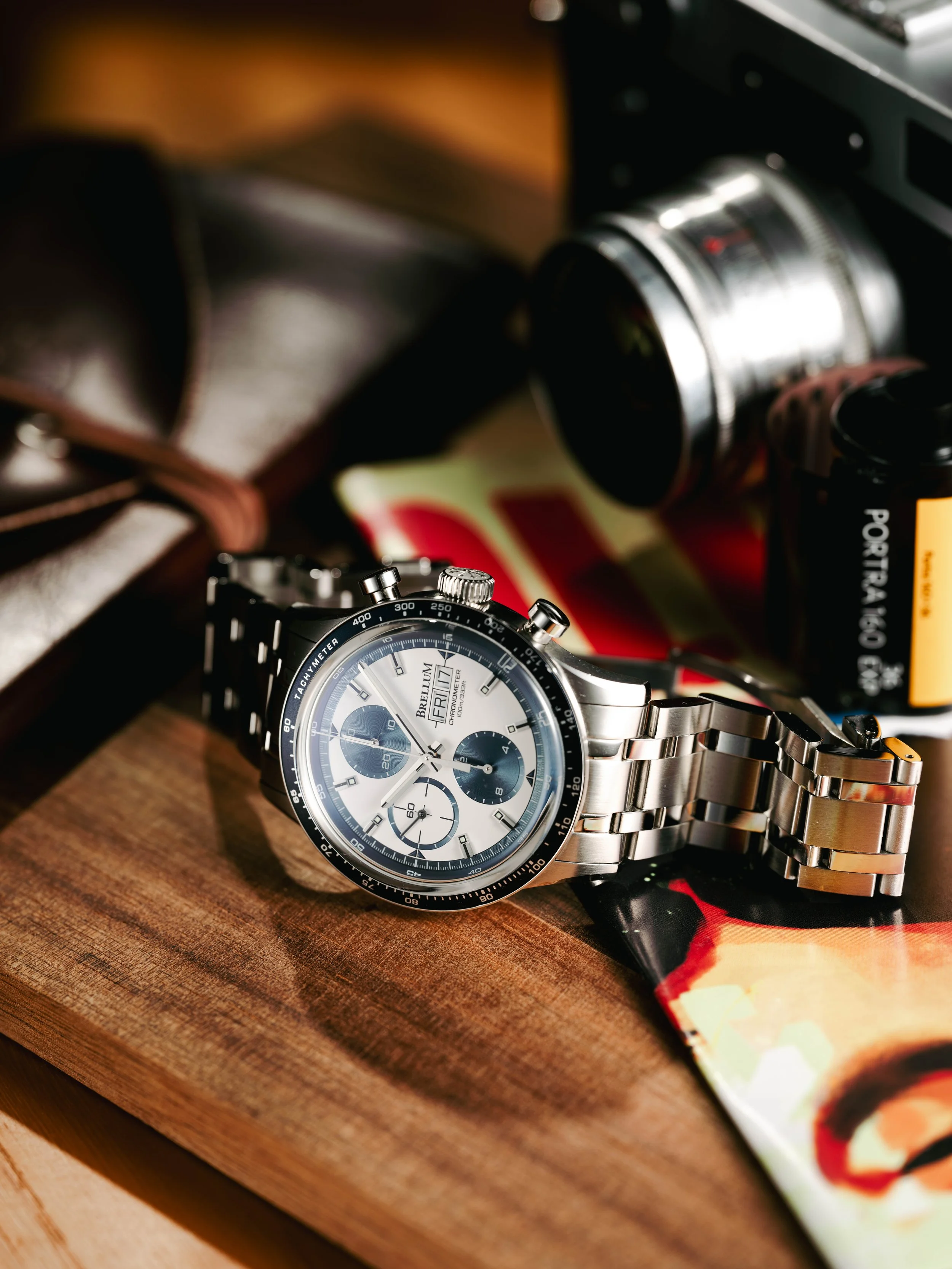 Silver men's wristwatch with a chronograph face, placed on a wooden surface among a magazine, a camera, and a can of beverage.