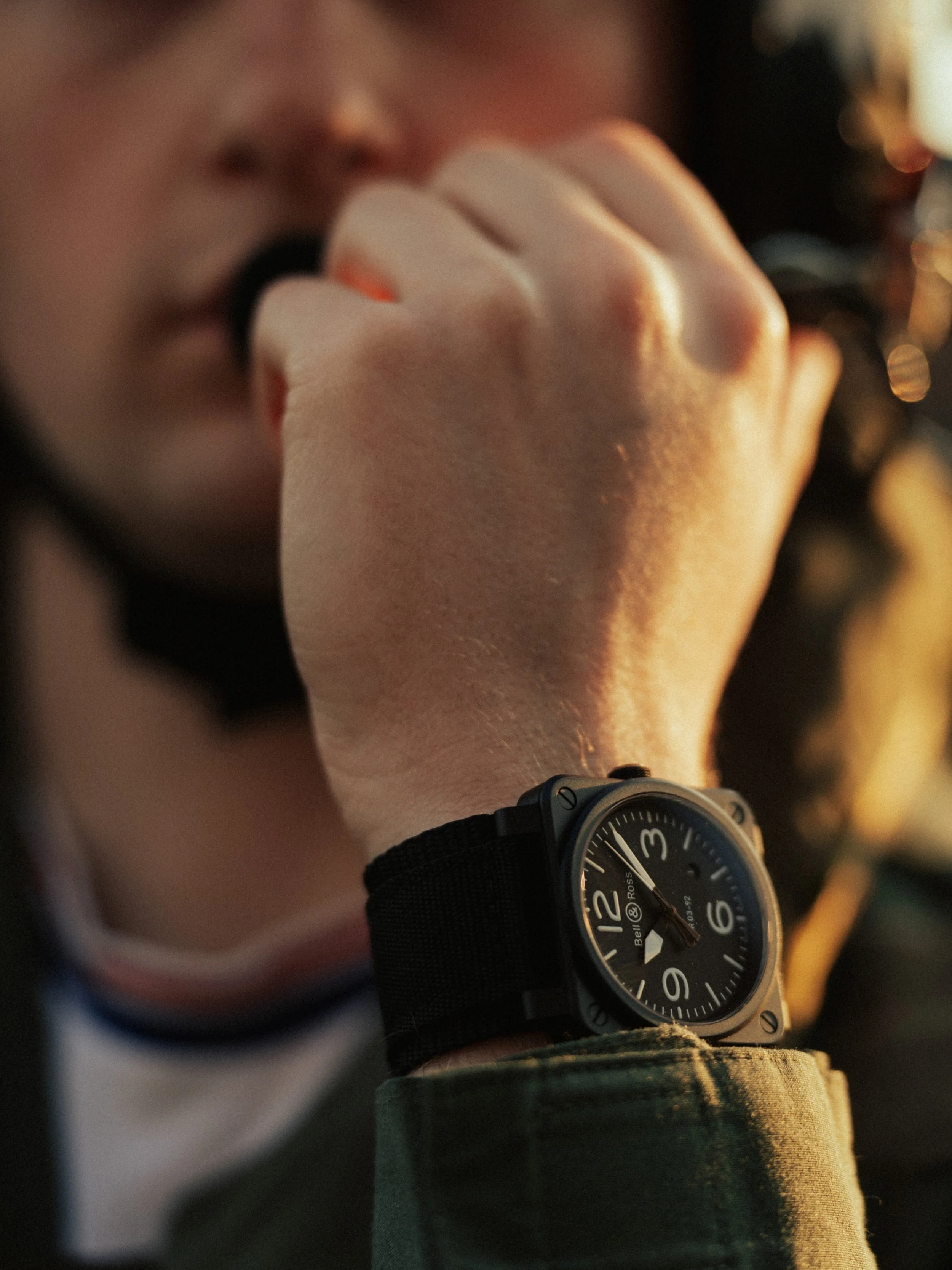 Person wearing a black Bell & Ross watch, holding a black object near their mouth with a blurred face and hand, wearing a green jacket.