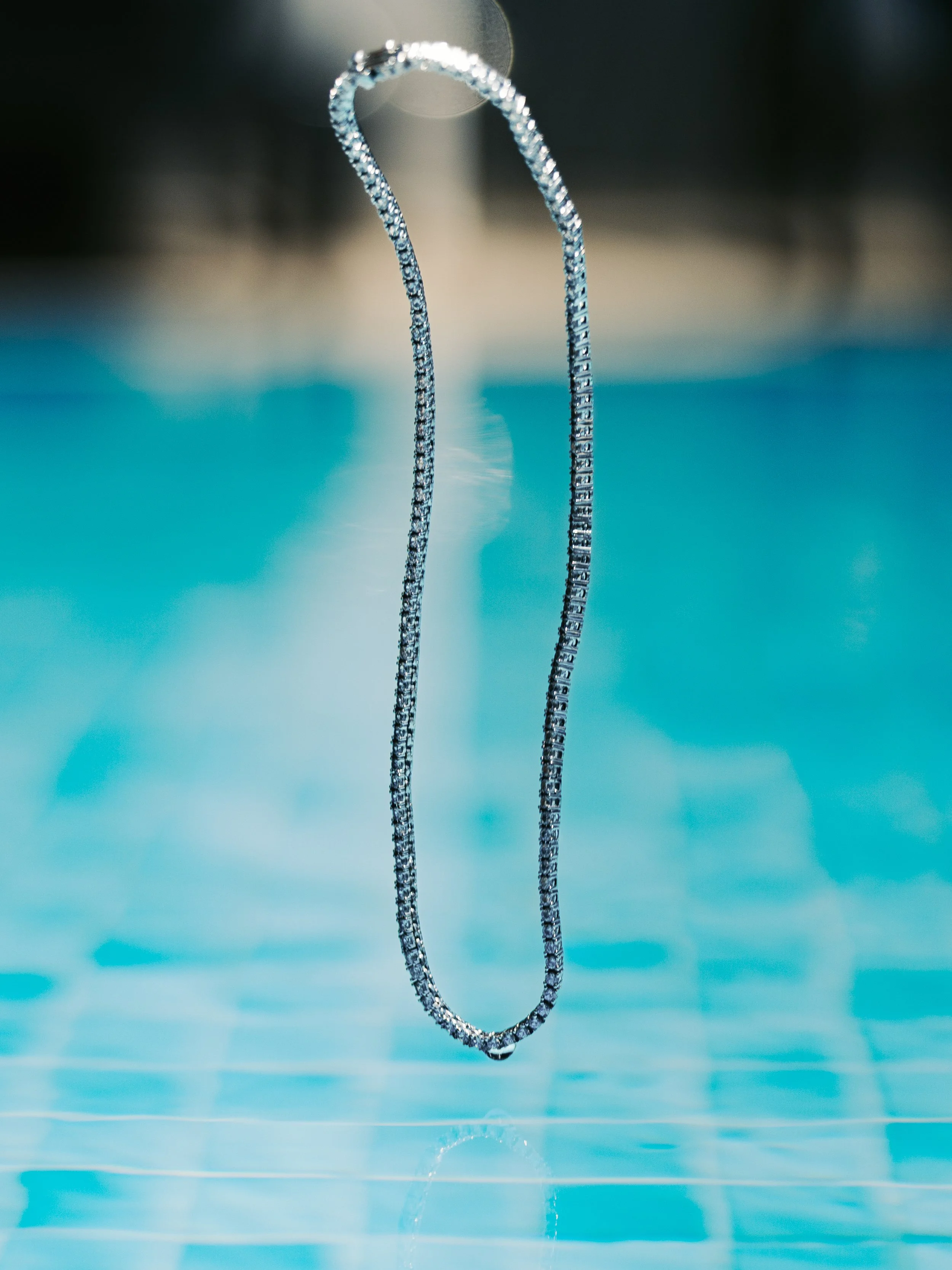 A close-up of a silver chain necklace hanging above a swimming pool with blue water.