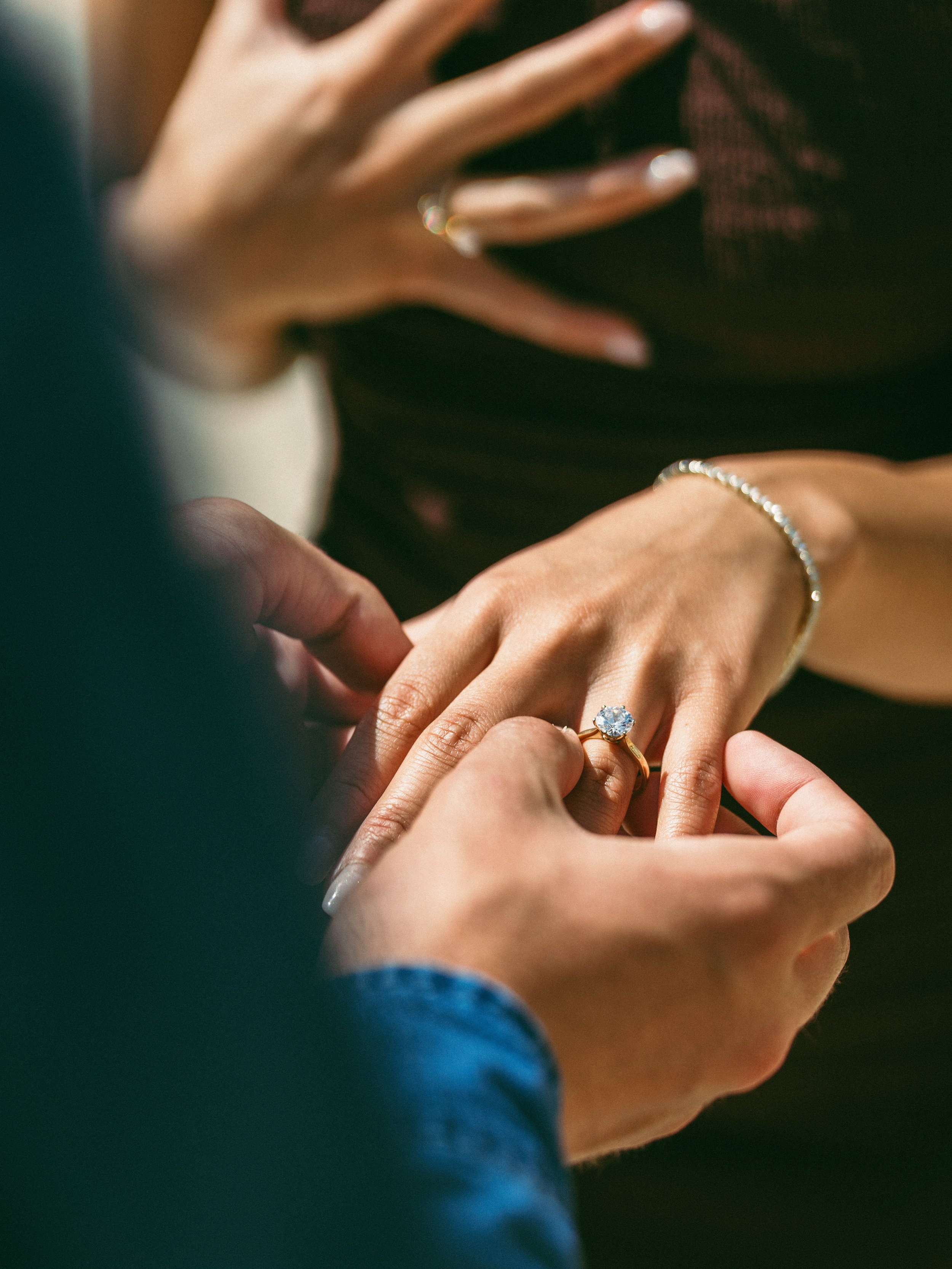 A person is being fitted with a ring during a wedding ceremony. The focus is on the woman's hand with a large gemstone ring, a bracelet, and a ring finger being helped by another person.