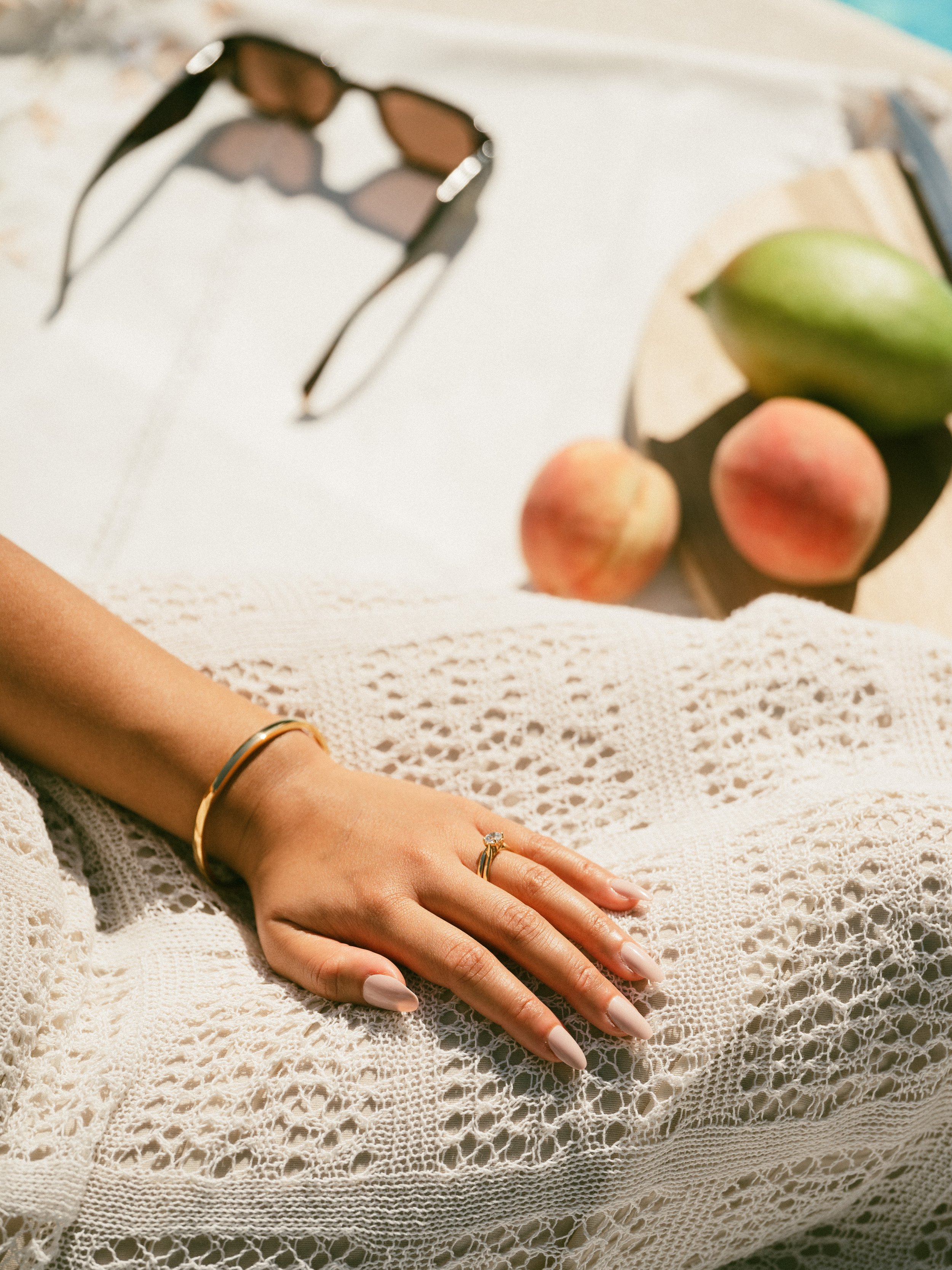 A woman's hand resting on a lace cloth, wearing a gold bracelet and a ring. In the background, sunglasses, a peach, a plum, and a book.