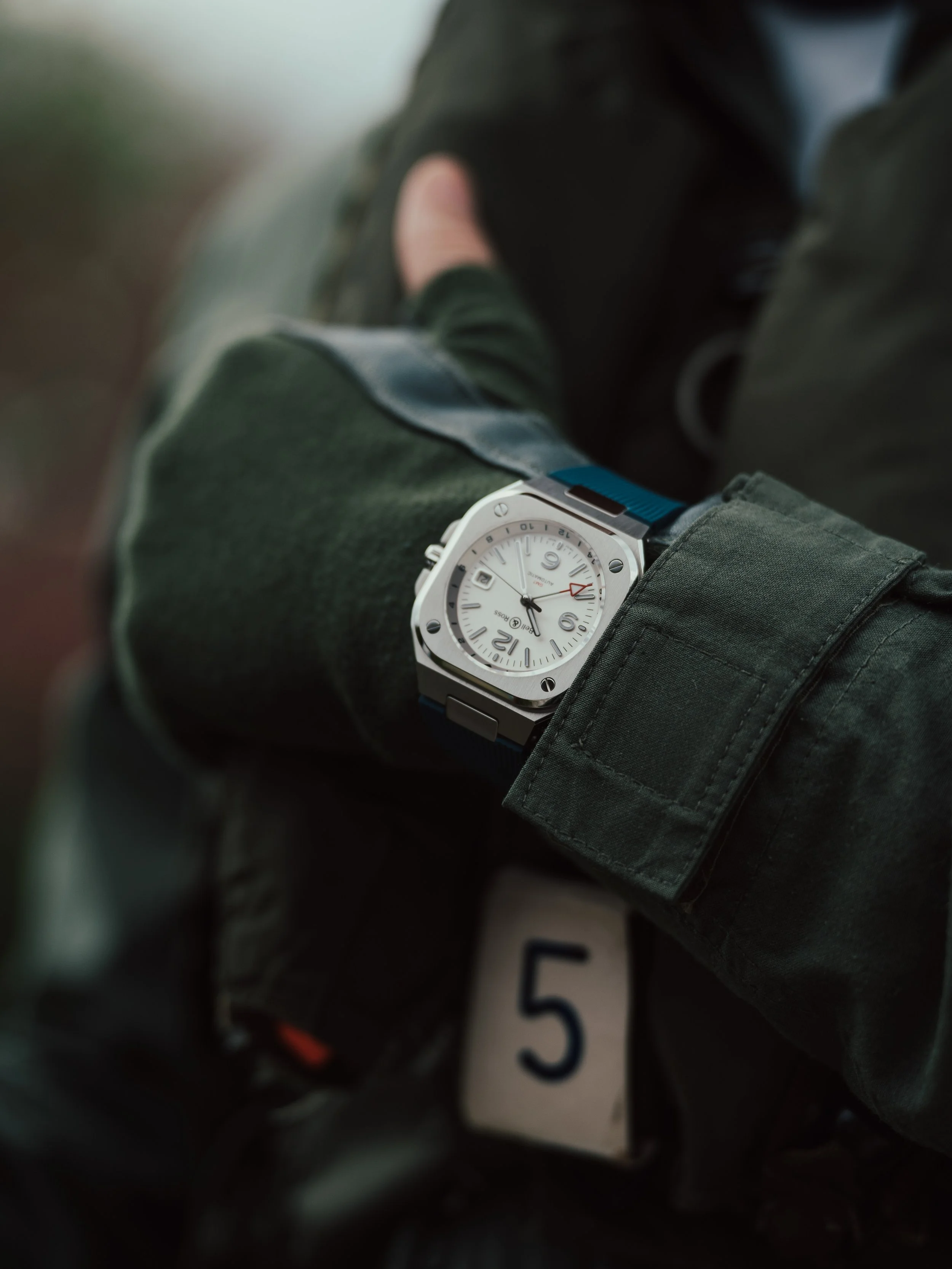 Close-up of a person's wrist wearing a silver watch with a white dial and black numerals, on a green jacket sleeve.