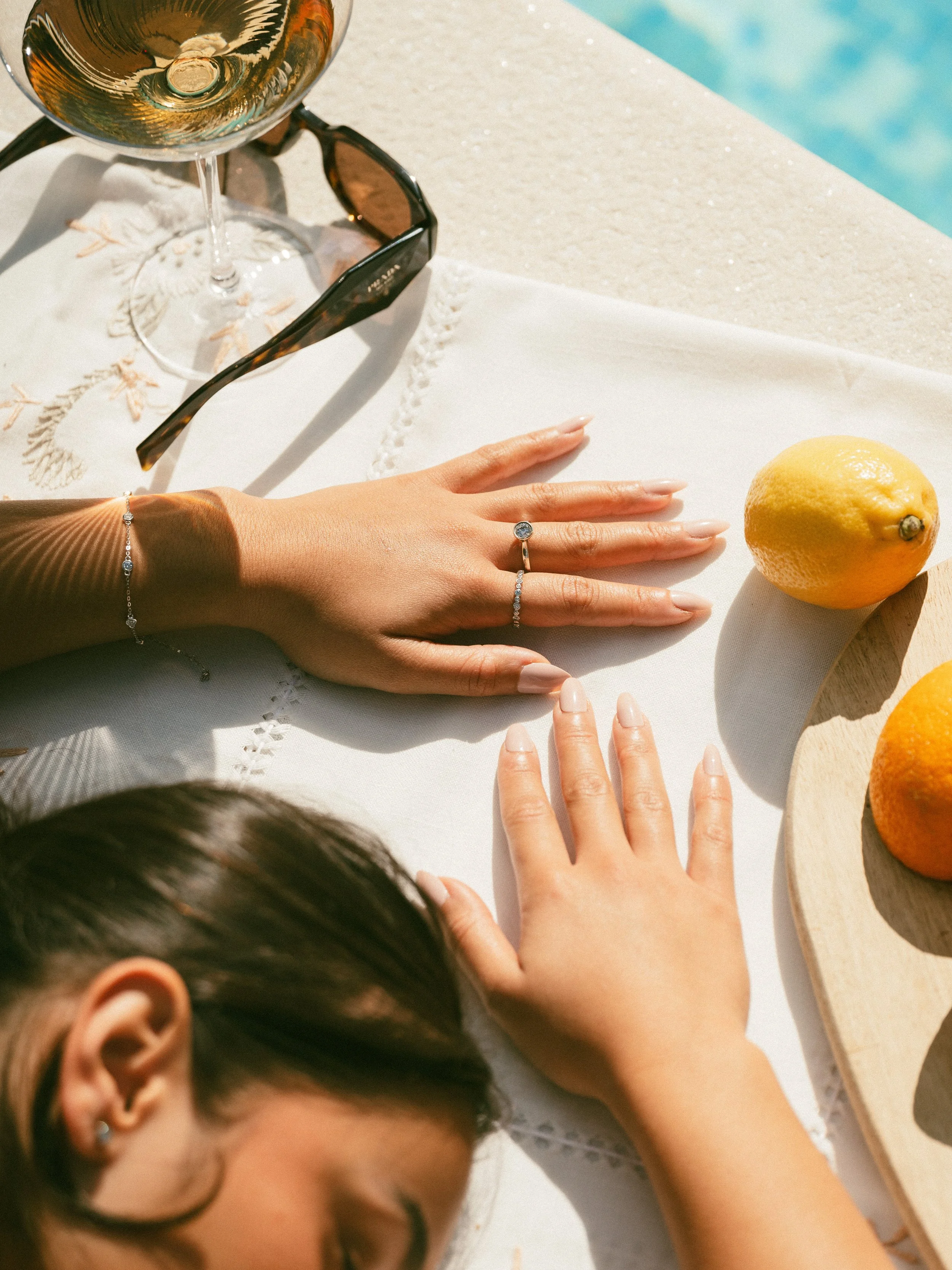 A woman's hand with rings and a bracelet resting on a white tablecloth at an outdoor setting with a lemon, orange, sunglasses, and a glass of wine nearby.