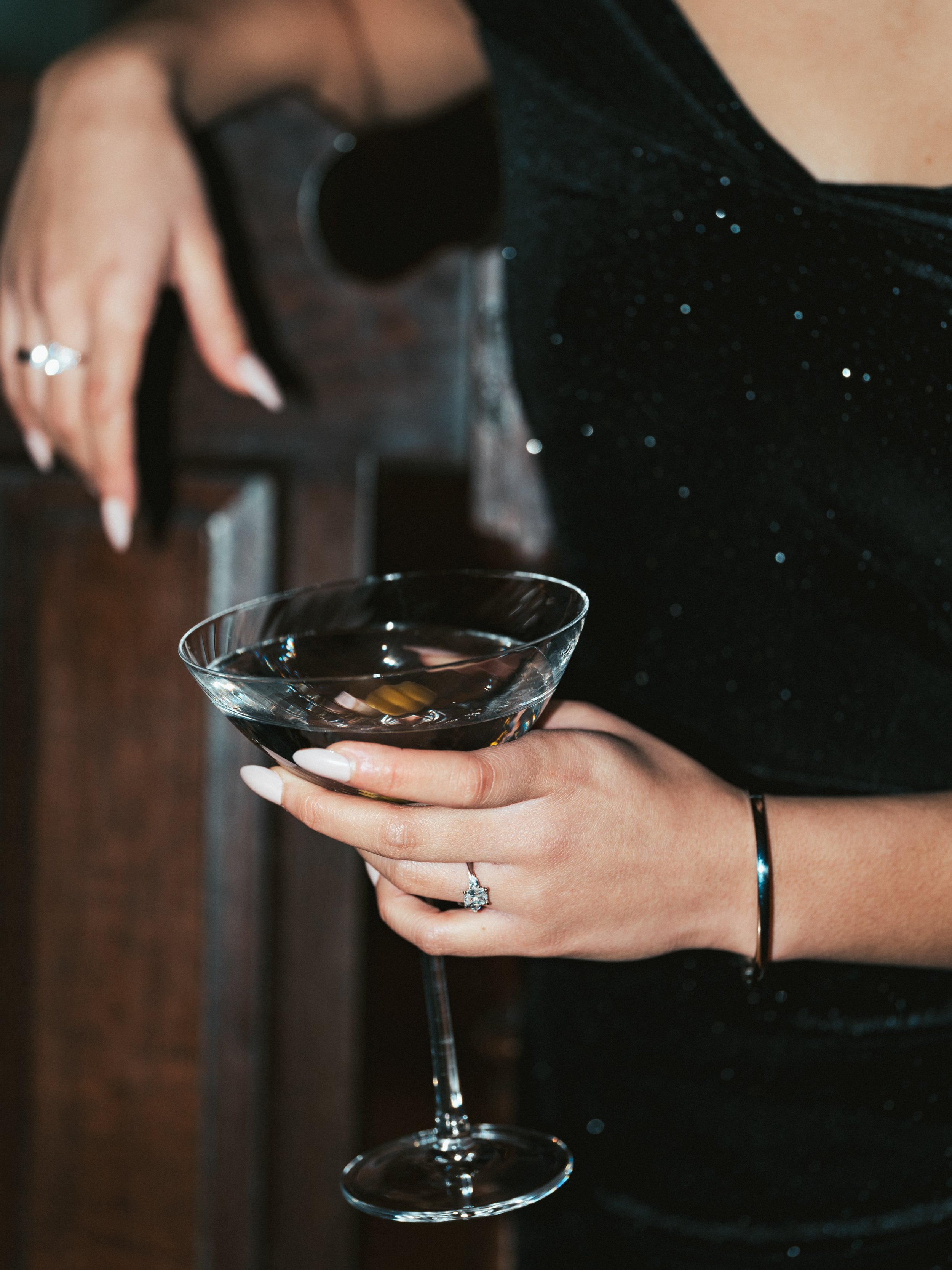 A woman holding a martini glass with a dark drink, wearing a black glittery dress, with a ring on her finger and a bracelet on her wrist.