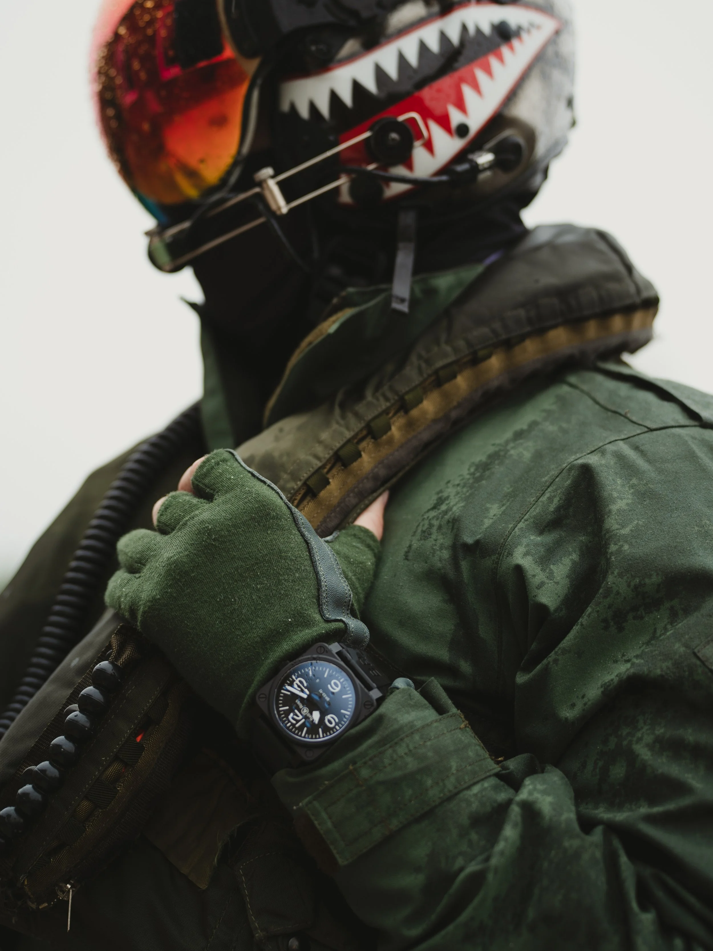 Close-up of a person wearing a camouflage jacket, a black watch, green gloves, and a helmet with a shark face design and reflective goggles.