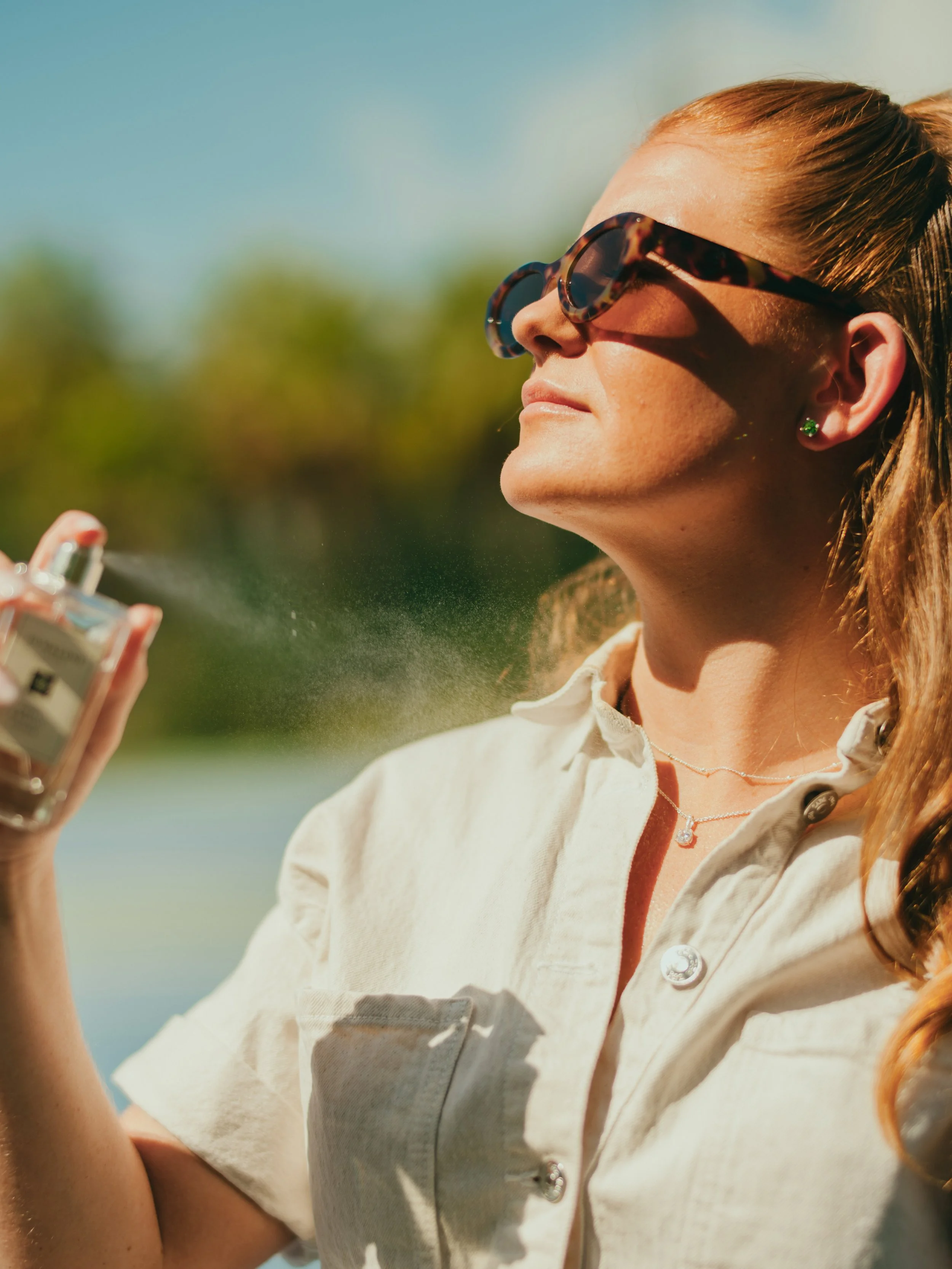 A woman wearing sunglasses sprays perfume outdoors.