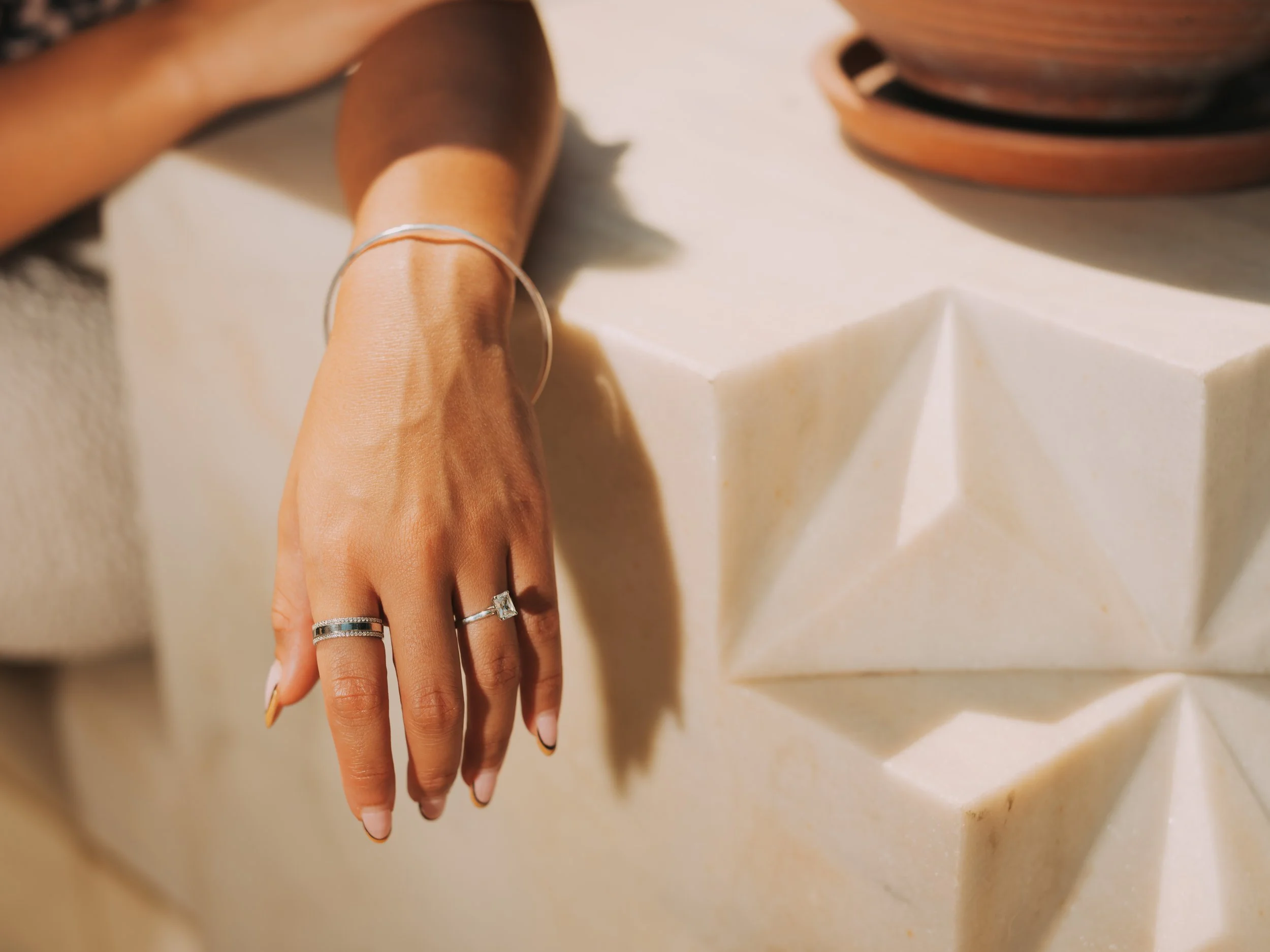 A hand with rings resting on a geometric white stone or sculpture, with wooden bowls or containers in the background.