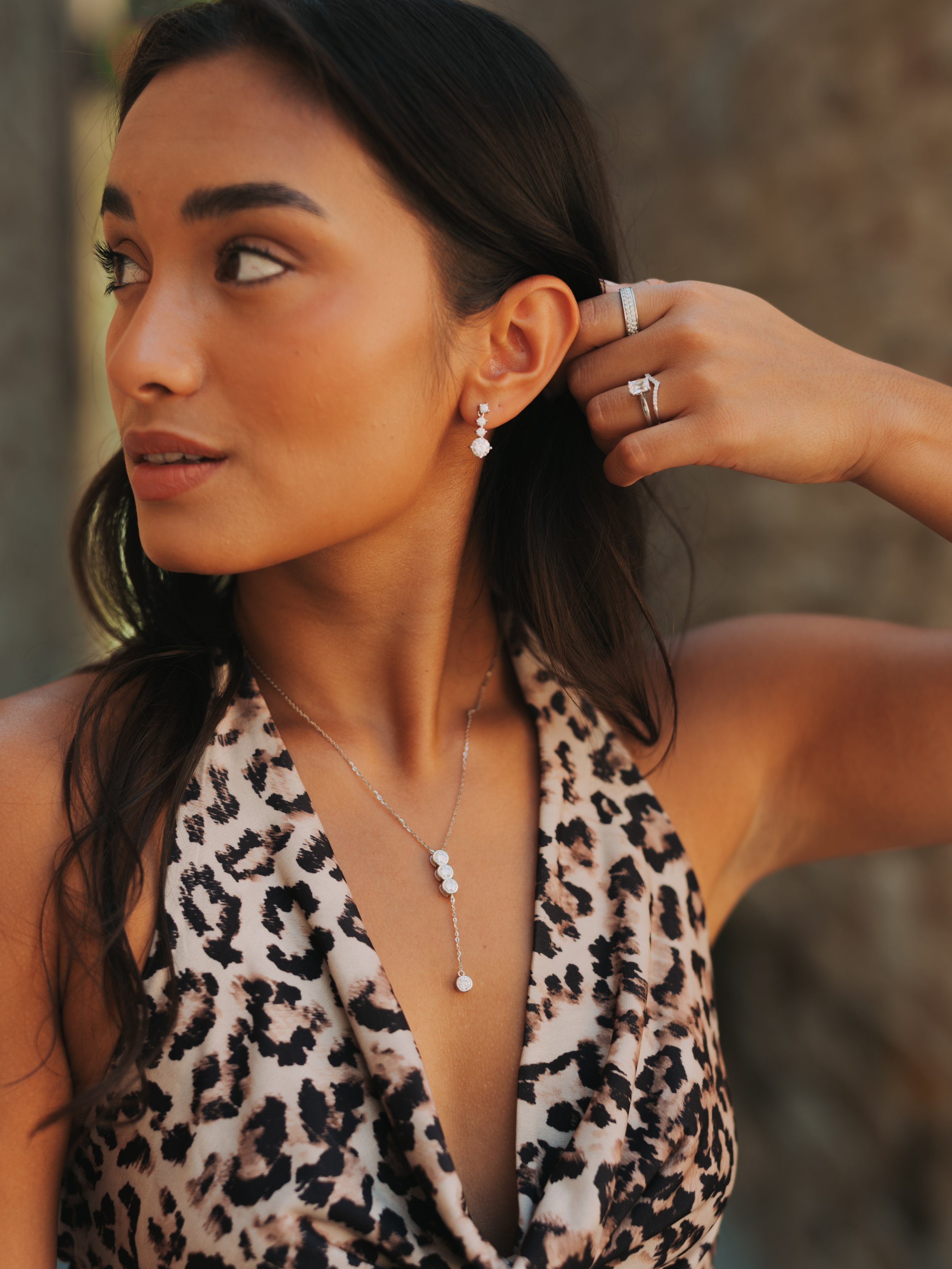 A woman with dark hair wearing diamond jewelry, including earrings, a necklace, and a ring, in a sleeveless leopard print top, adjusting her earring outdoors.