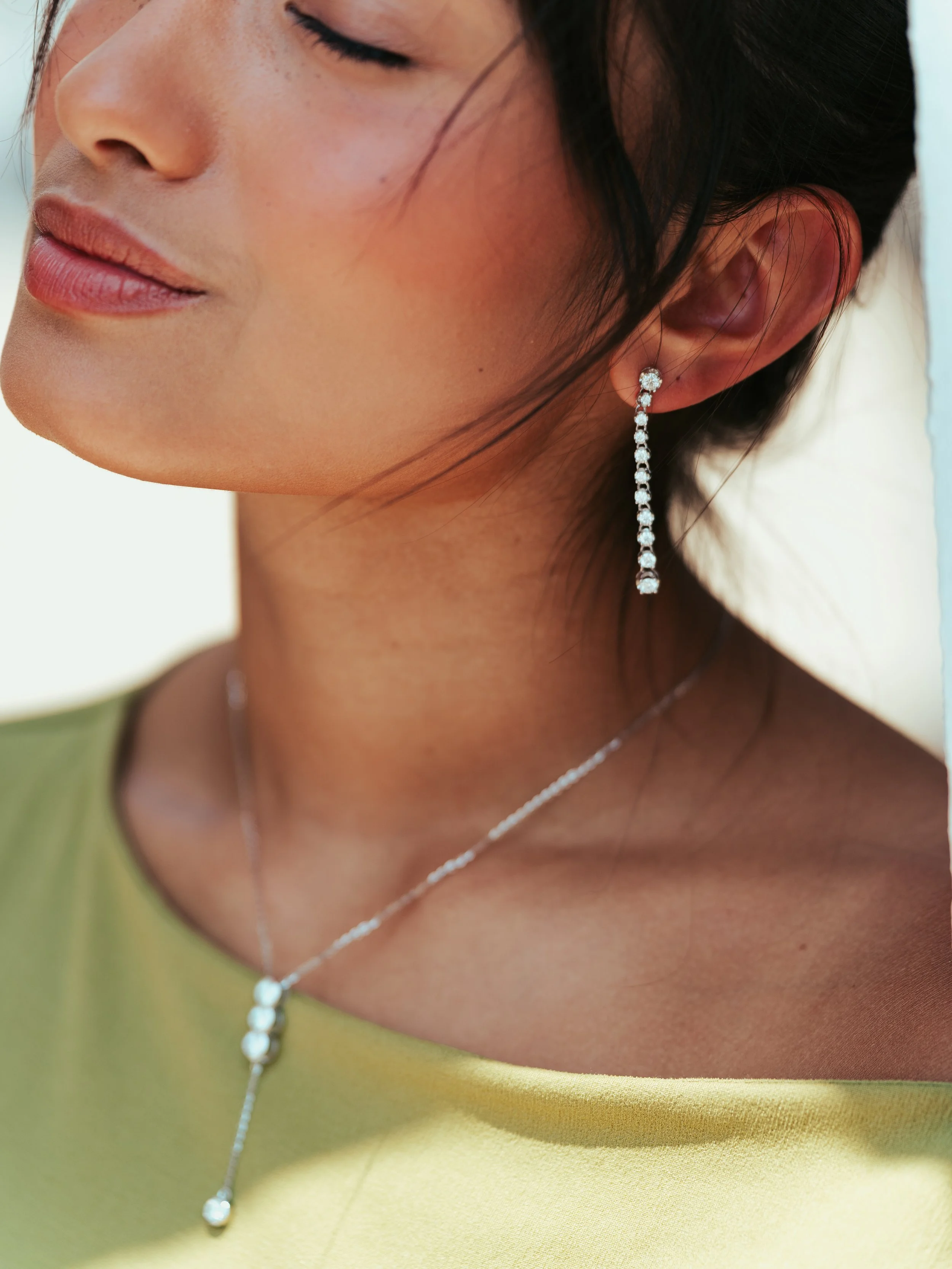 Close-up of a woman's face and neck showing jewelry, including long dangling diamond earrings and a matching necklace with diamonds, against a blurred background.