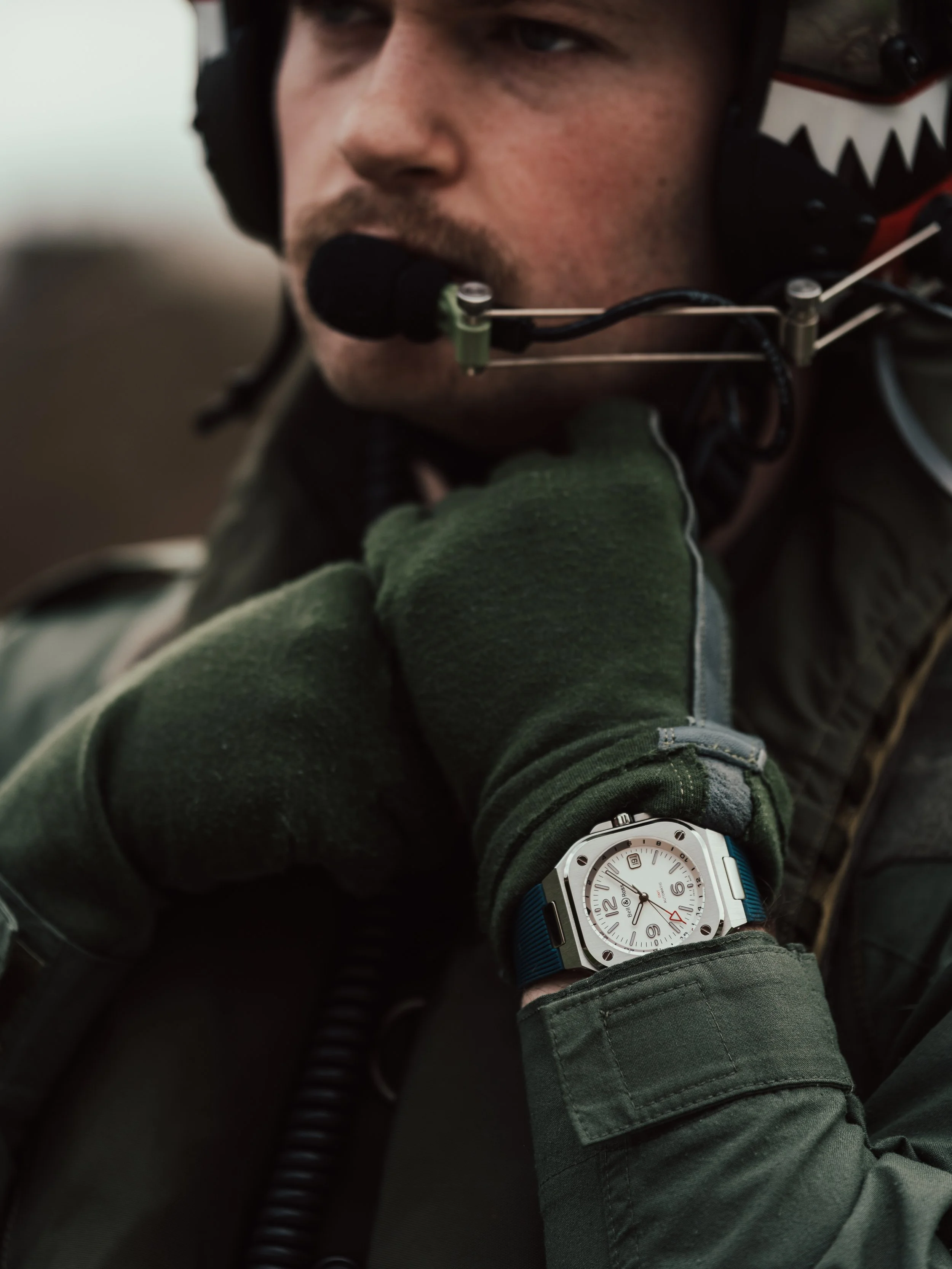 A man wearing a military-style jacket, green gloves, a pilot's headset, and a watch on his wrist, looking to the side.