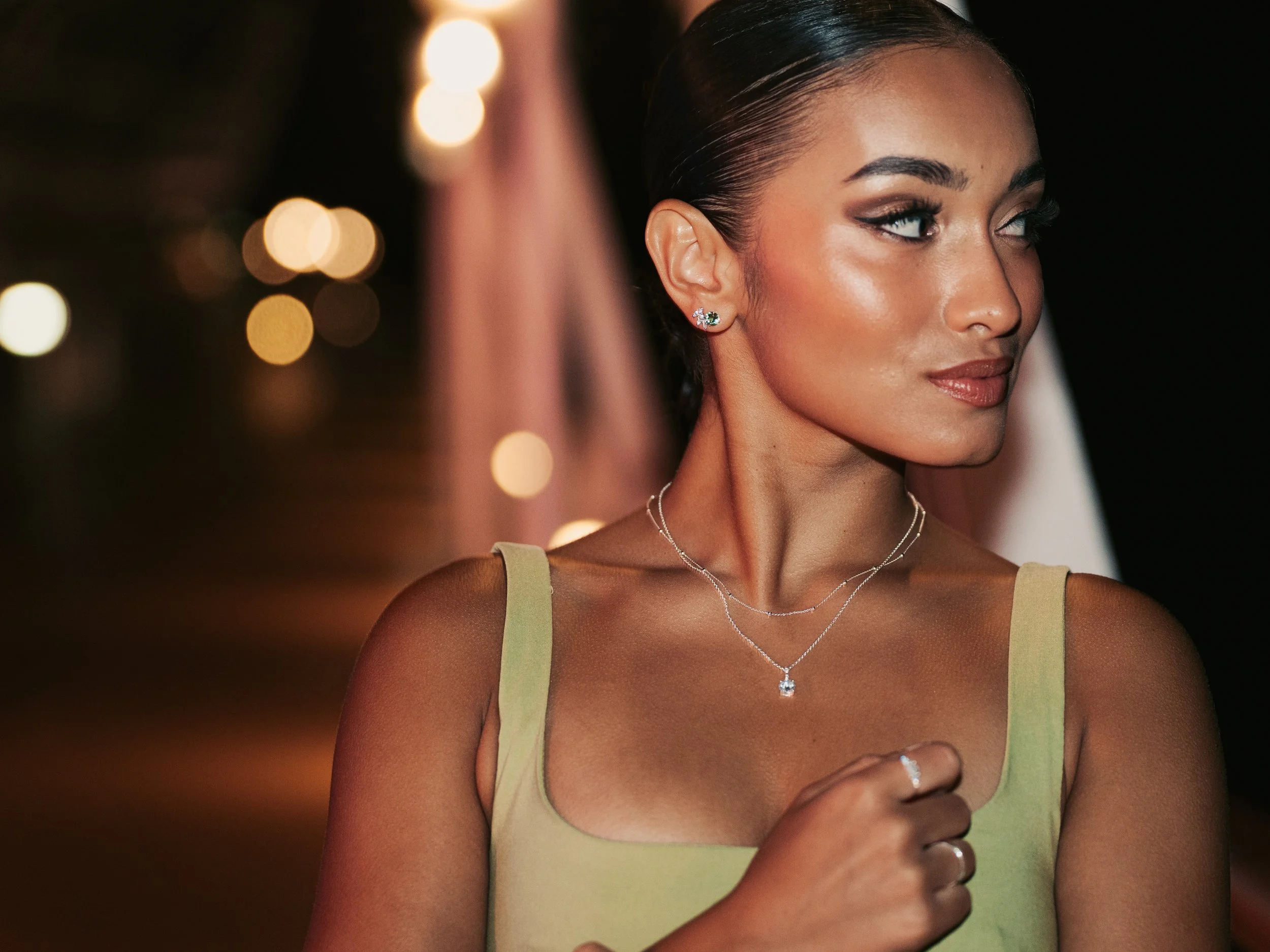 A woman with dark hair slicked back, wearing a sleeveless yellow-green top, silver jewelry, and eye makeup, is looking to her right at night with bokeh lights in the background.