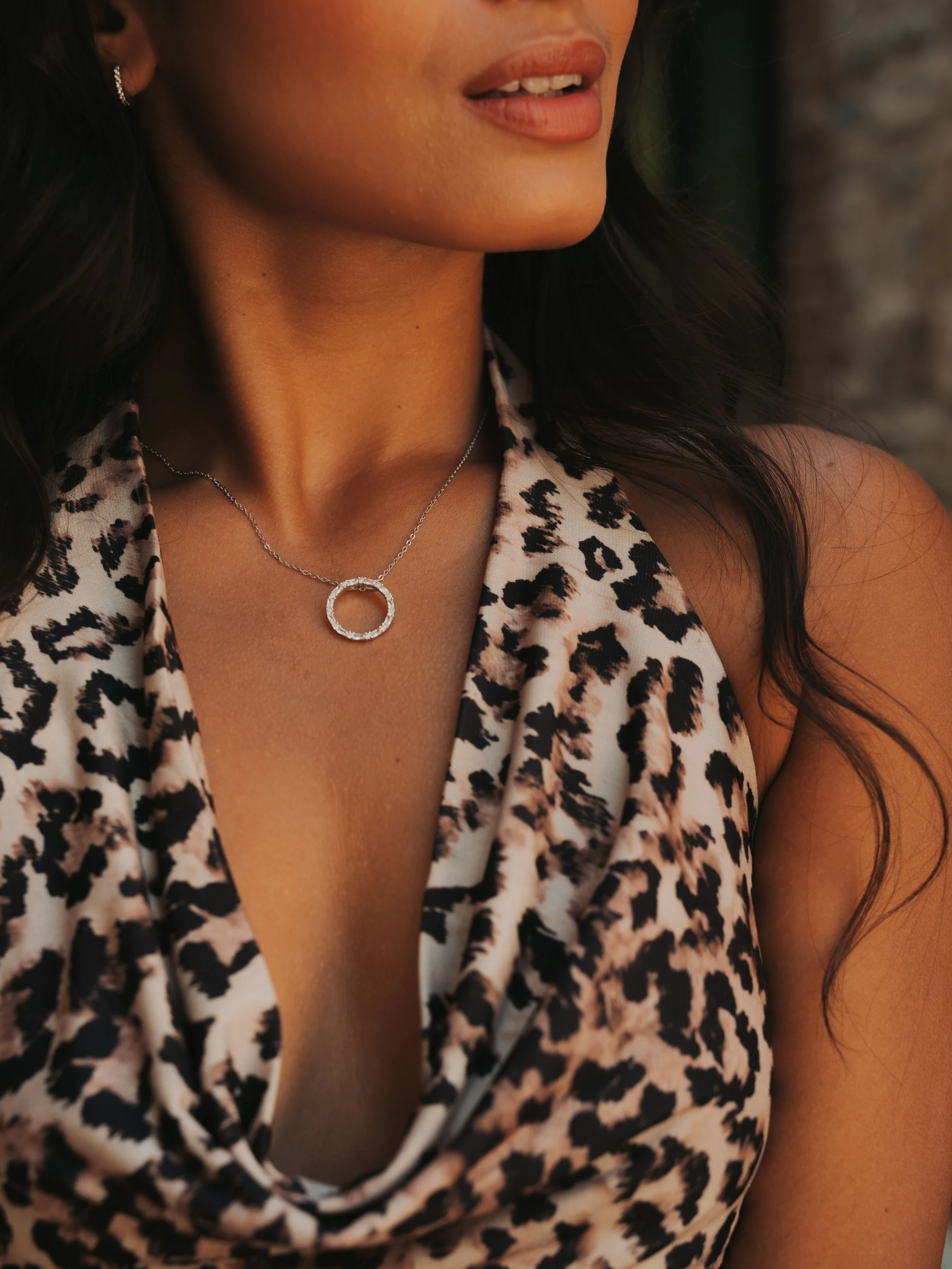 Close-up of a woman's neck and upper chest showing a silver necklace with a circular, textured pendant, wearing a leopard print sleeveless top with a cowl neckline.