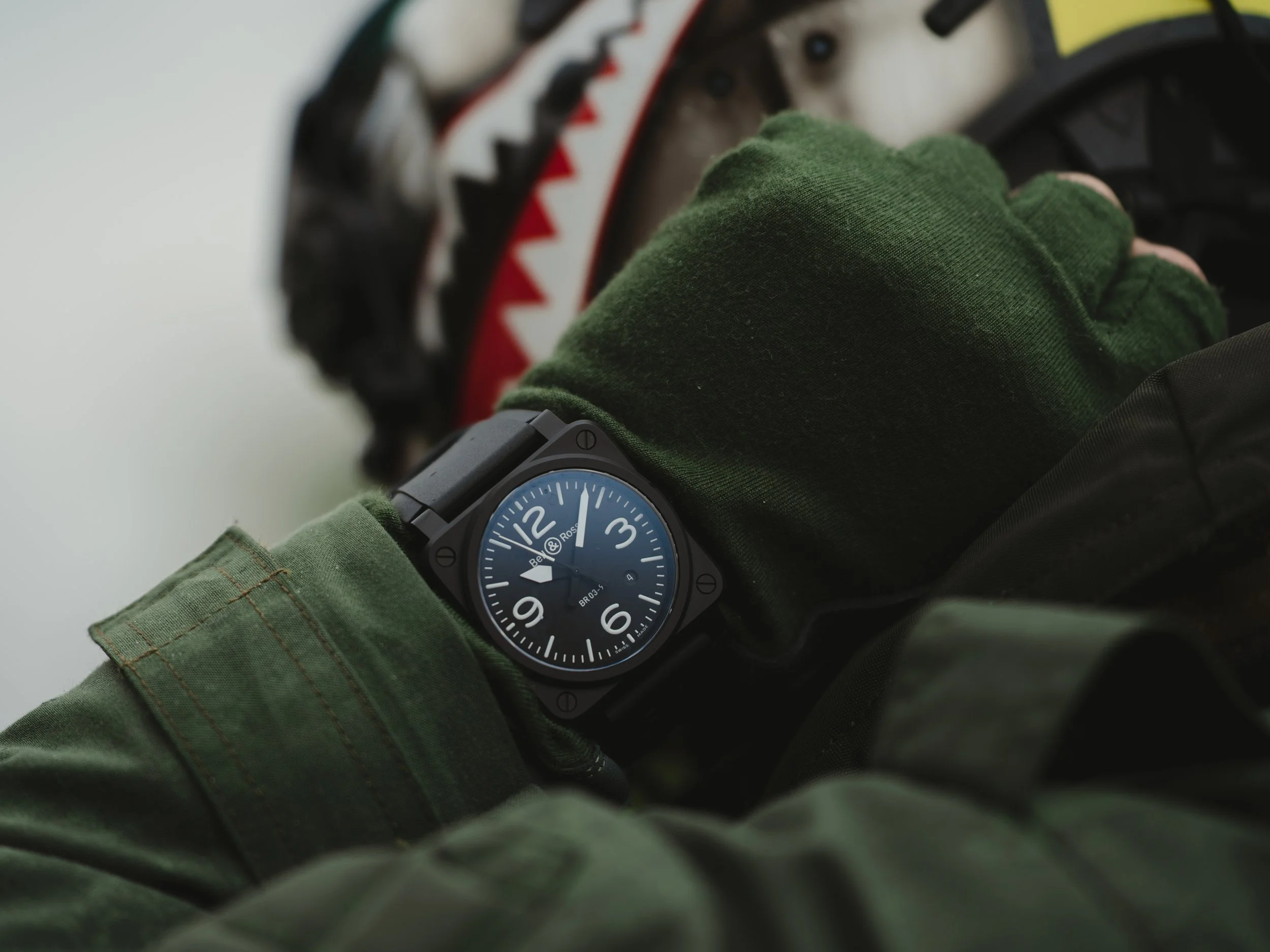 A person's wrist wearing a black Bell & Ross watch with a black strap, showing the time as 11:52, with a green gloved hand adjusting a red and black bicycle gear shifter.