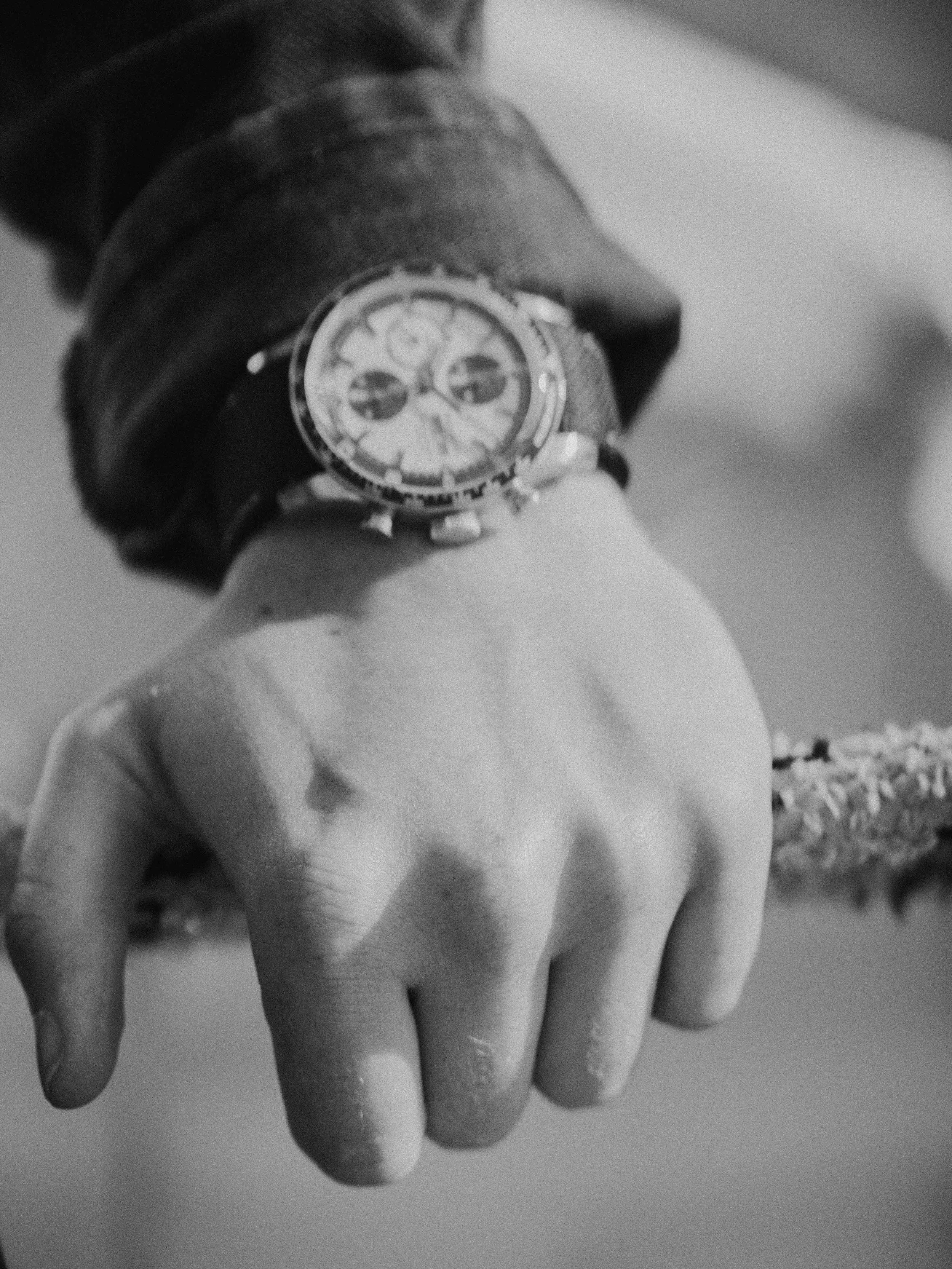 Close-up of a person's wrist wearing a large wristwatch with a dark strap, and their hand hanging down with fingers slightly curled. The person is wearing a textured long-sleeve garment.