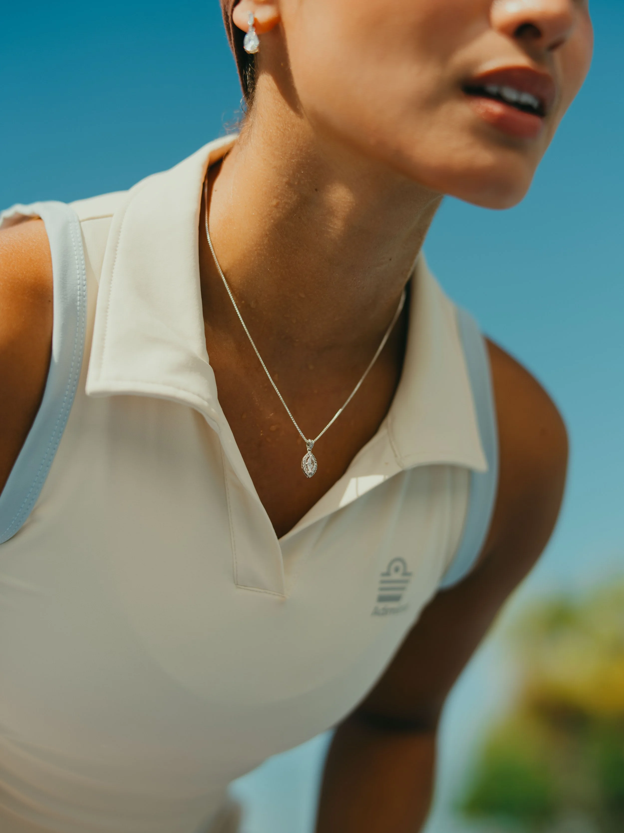 Close-up of a woman wearing a white athletic top with a silver necklace and earrings, outdoors in bright sunlight, with a clear blue sky in the background.