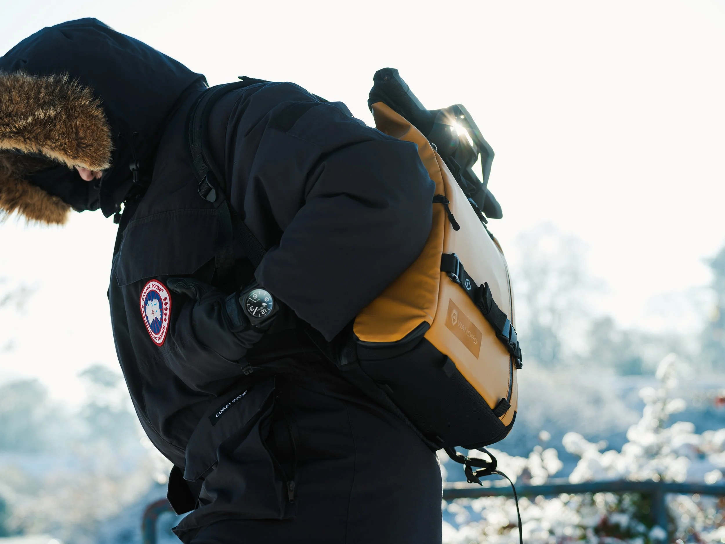 A person dressed in winter clothing, including a black jacket with a Canada Goose patch, is carrying a tan and black backpack with a logo, outdoors in a snowy environment.