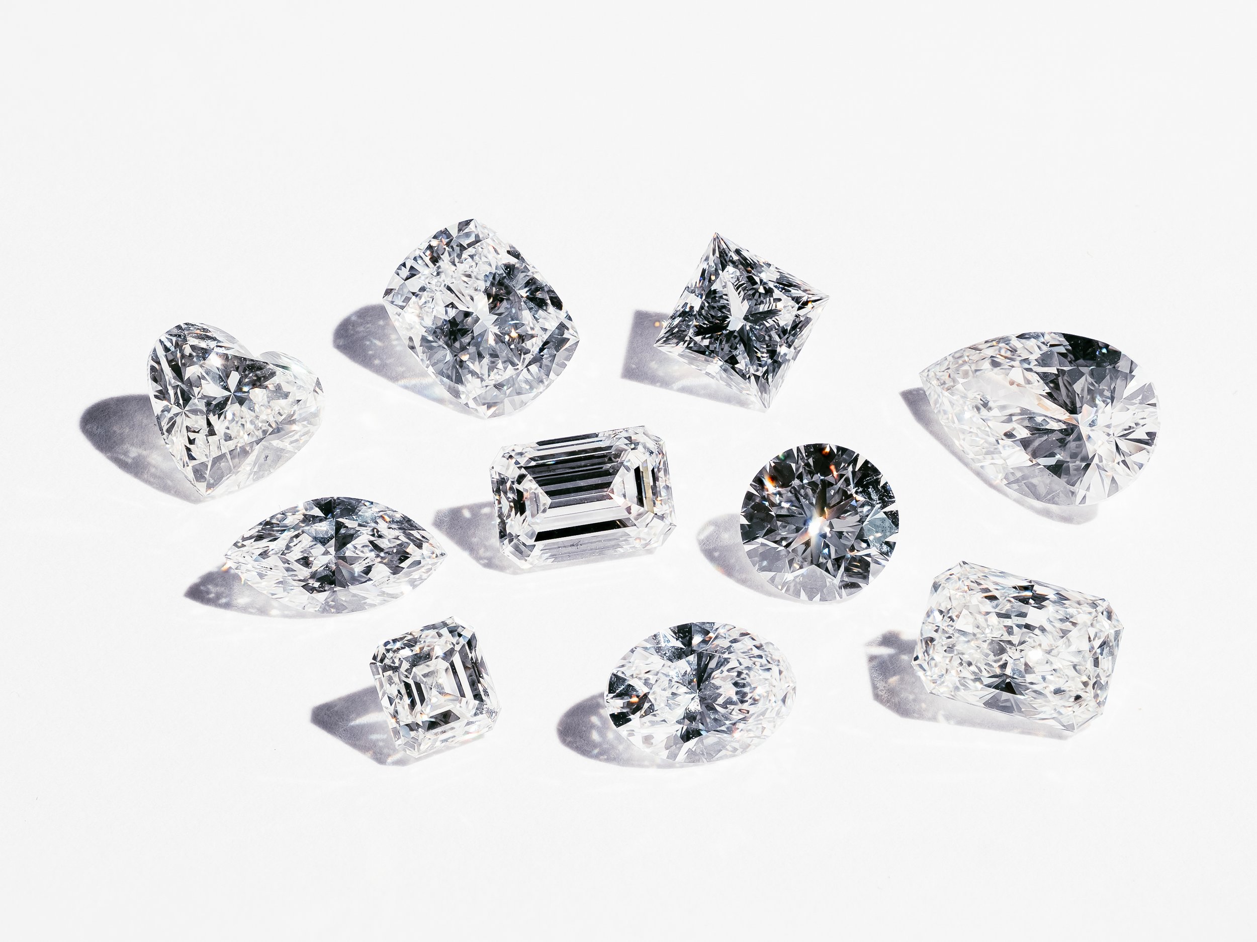 A collection of various cut diamonds with different shapes and sizes, placed on a white surface, casting shadows.