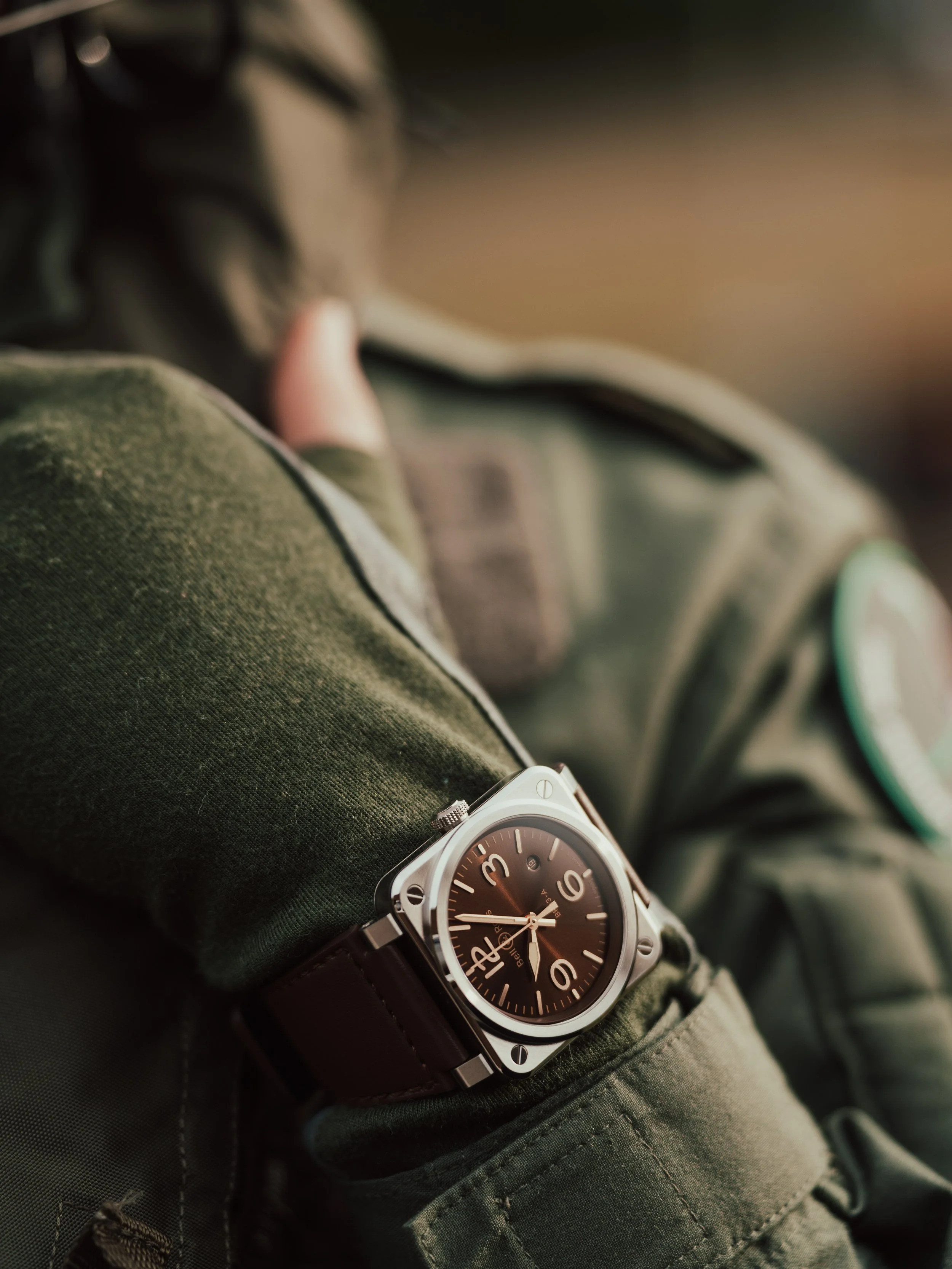 Close-up of a wristwatch with a square silver case and black dial, worn on a person's wrist with a green tactical jacket.