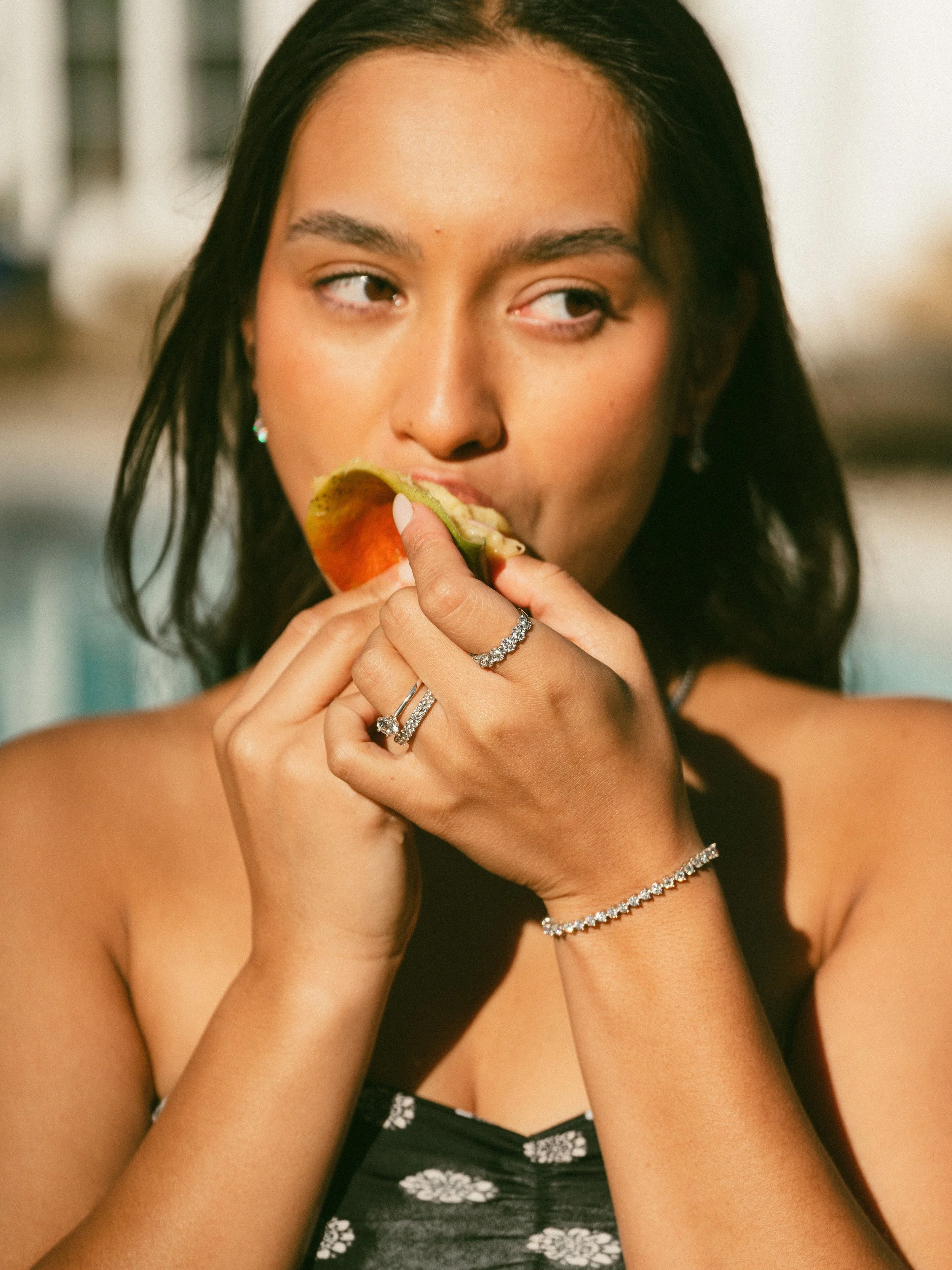 A woman with dark hair is eating a fruit, possibly an apple, while holding it with both hands. She is wearing rings and a diamond bracelet.