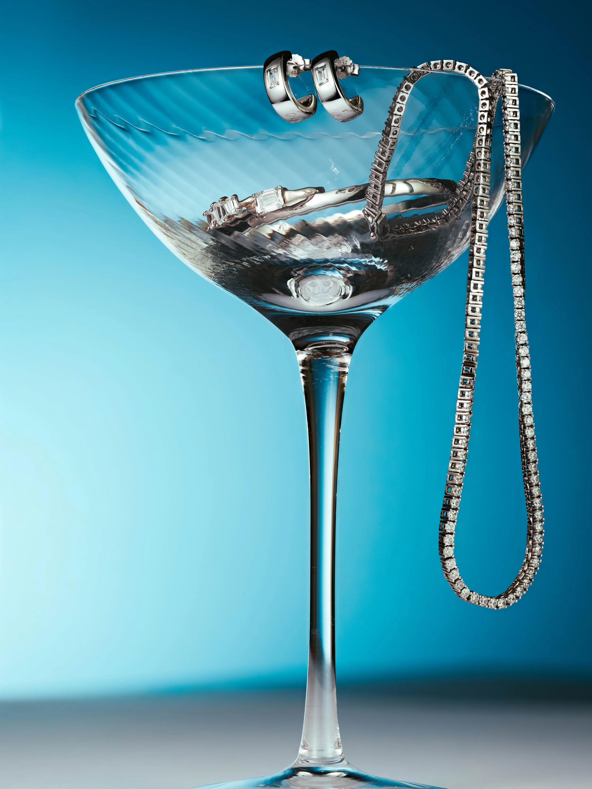 A martini glass with silver jewelry, including a chain necklace, bracelet, and earrings, hanging over the rim against a blue background.