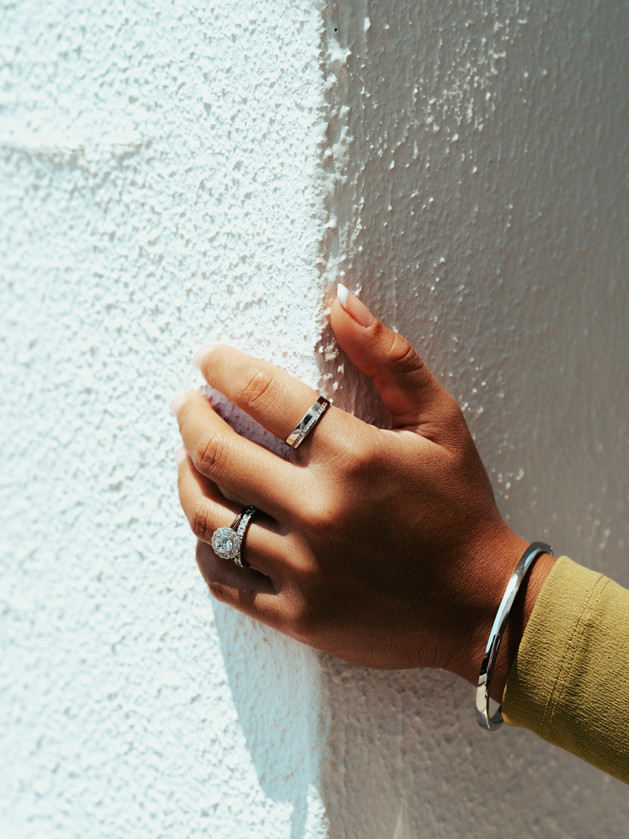A person's hand with rings and a bracelet touches a textured white wall.