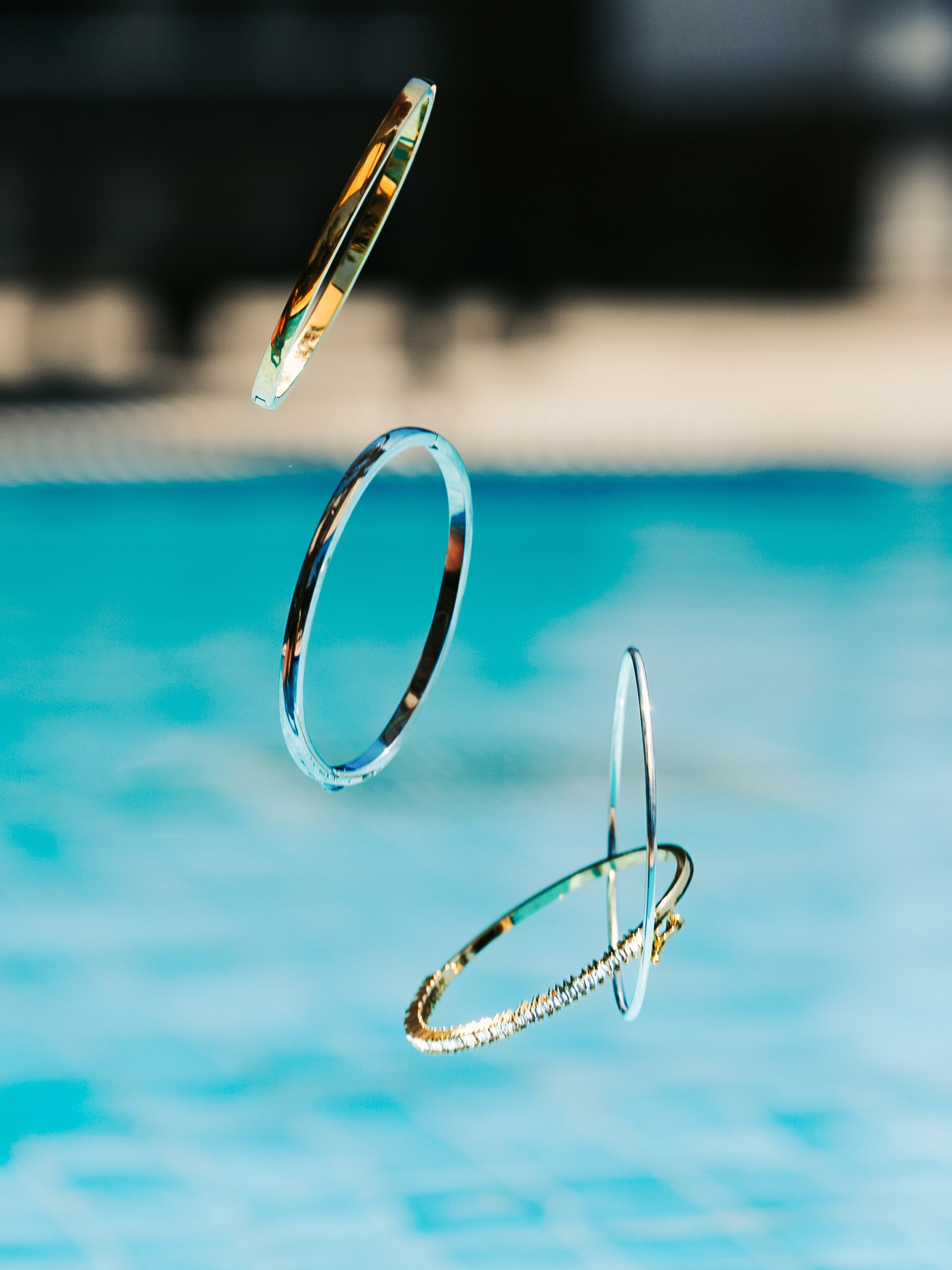 Three metallic bangles suspended in mid-air above a swimming pool with blue water.