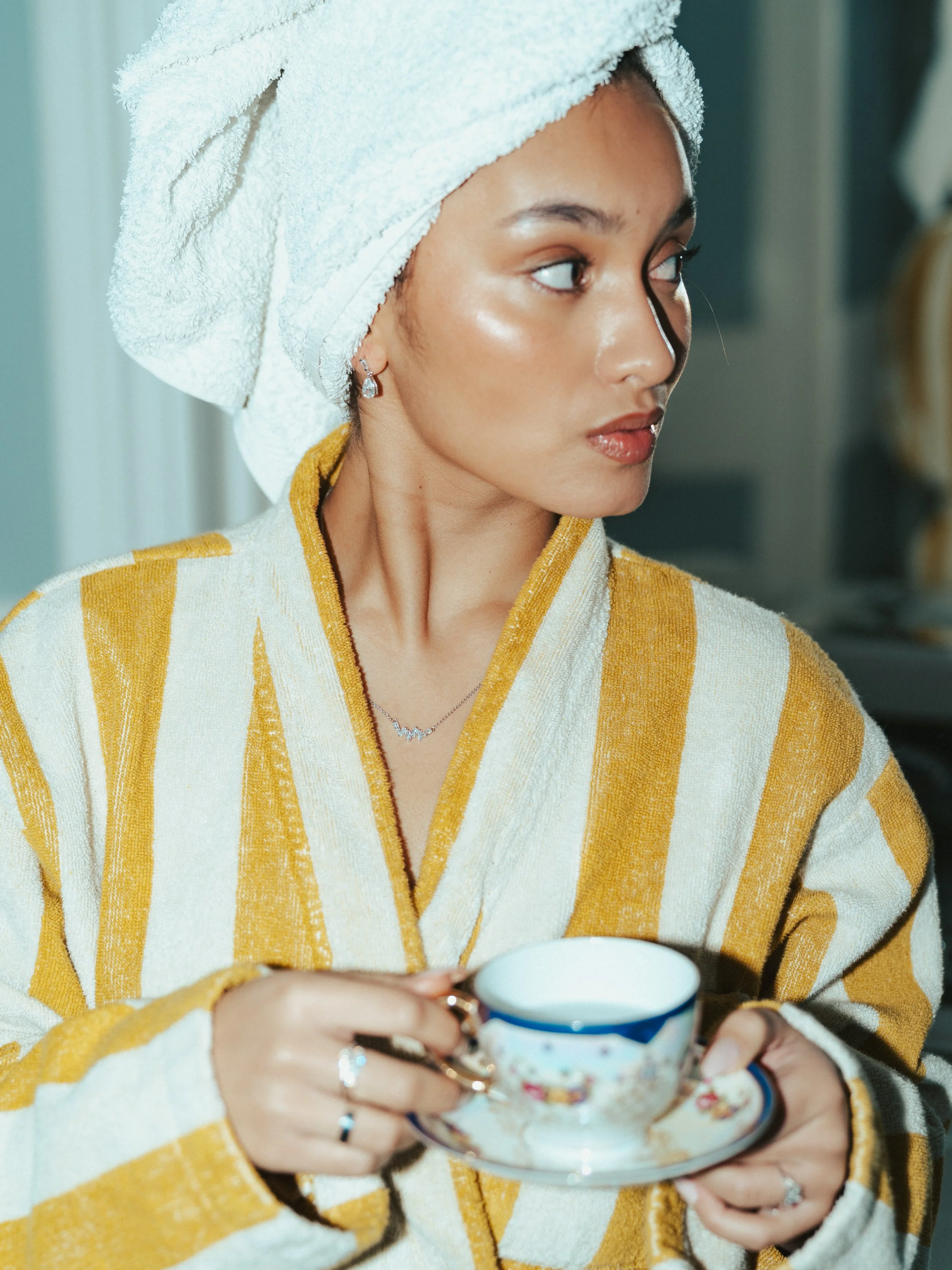A woman wrapped in a white towel with a towel turban on her head, holding a teacup and saucer, wearing jewelry, and looking to the right.