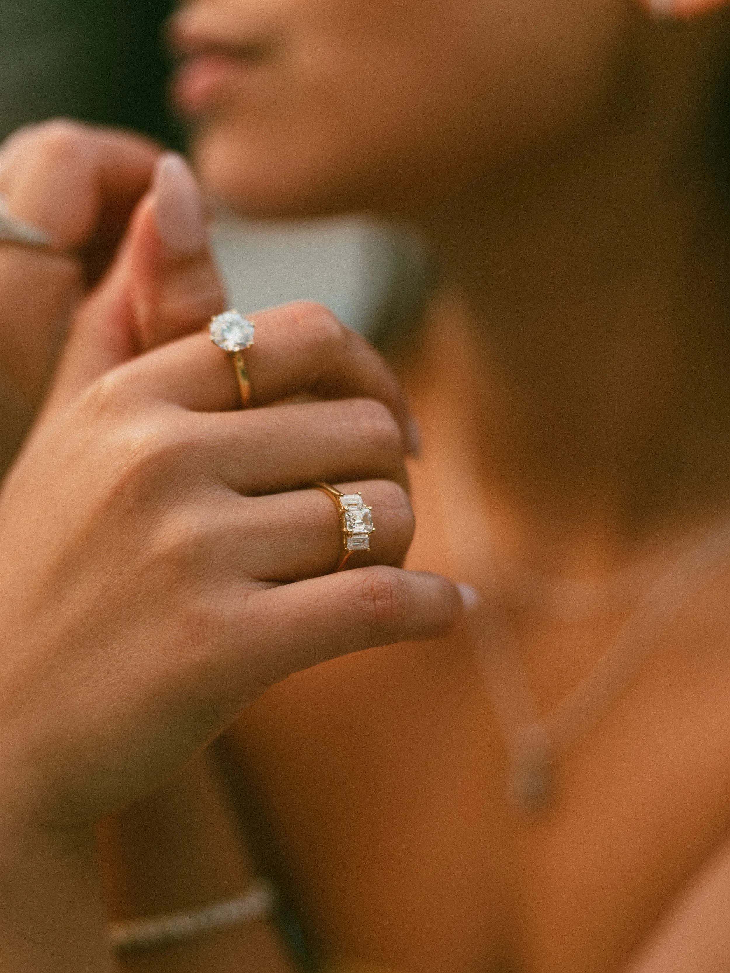 Close-up of a woman's hand wearing a gold ring with a large round diamond and a gold ring with three baguette diamonds, with blurred background of her face and shoulder.