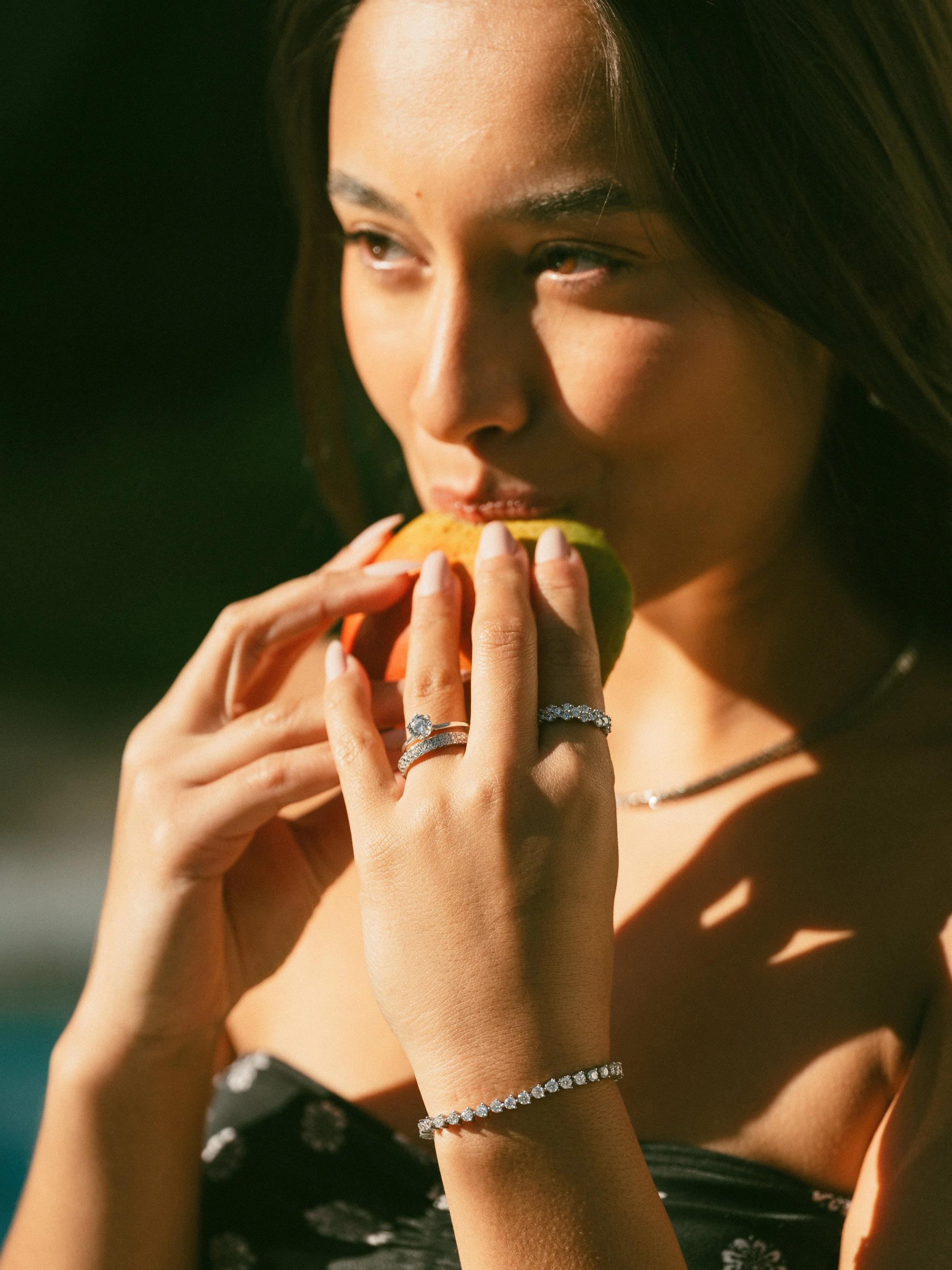 A woman with long dark hair eating a green apple, wearing a diamond ring, bracelet, and necklace.