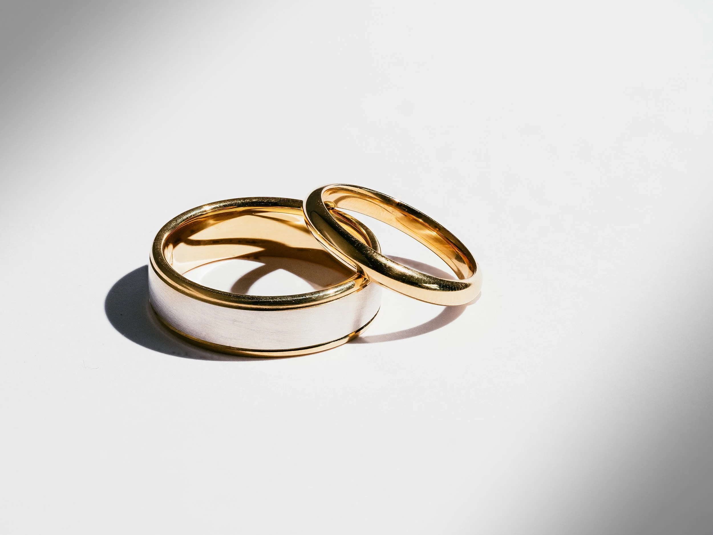 Close-up of two intertwined wedding rings, one silver and one gold, on a white background with shadows.