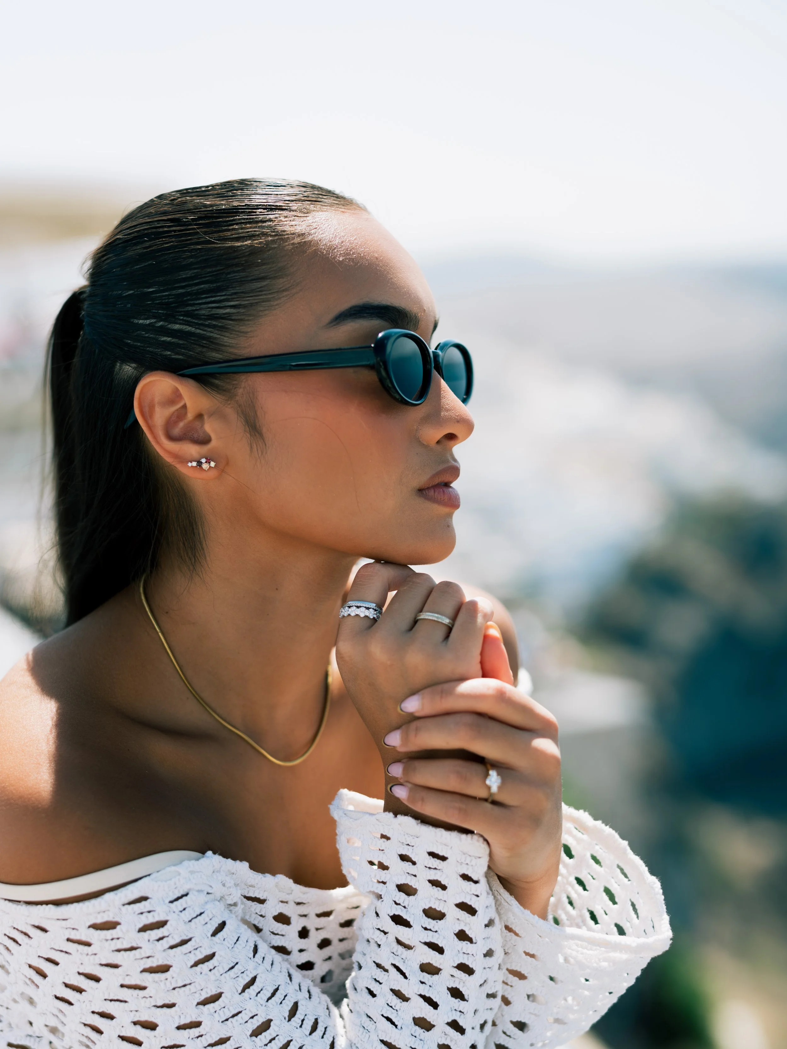 A woman with dark hair tied back, wearing black sunglasses, earrings, rings, and a gold necklace, dressed in a white crochet top, posing outdoors with a blurred ocean background.