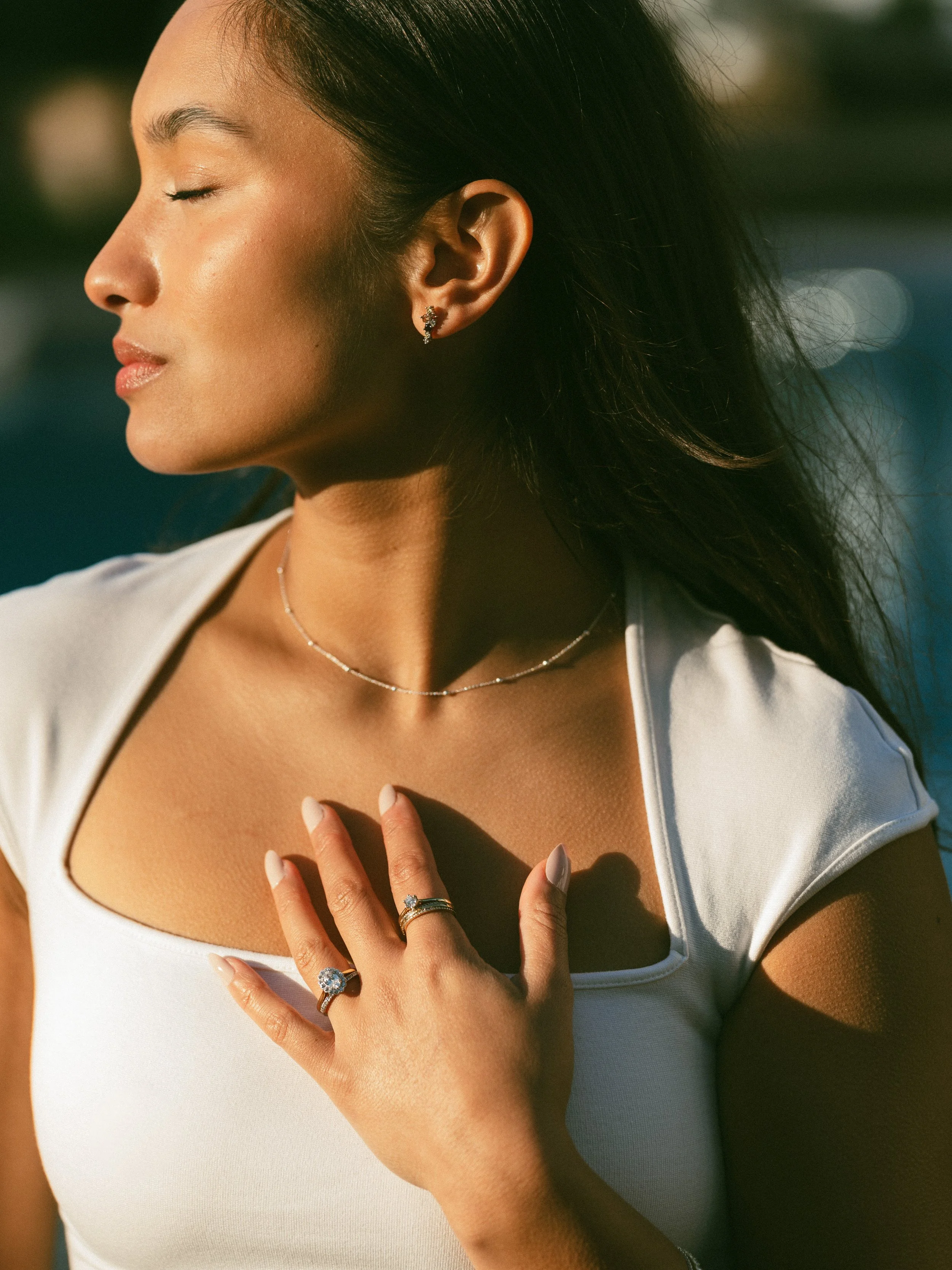 Close-up of a woman wearing jewelry, with her eyes closed, touching her chest, in a sunny outdoor setting.