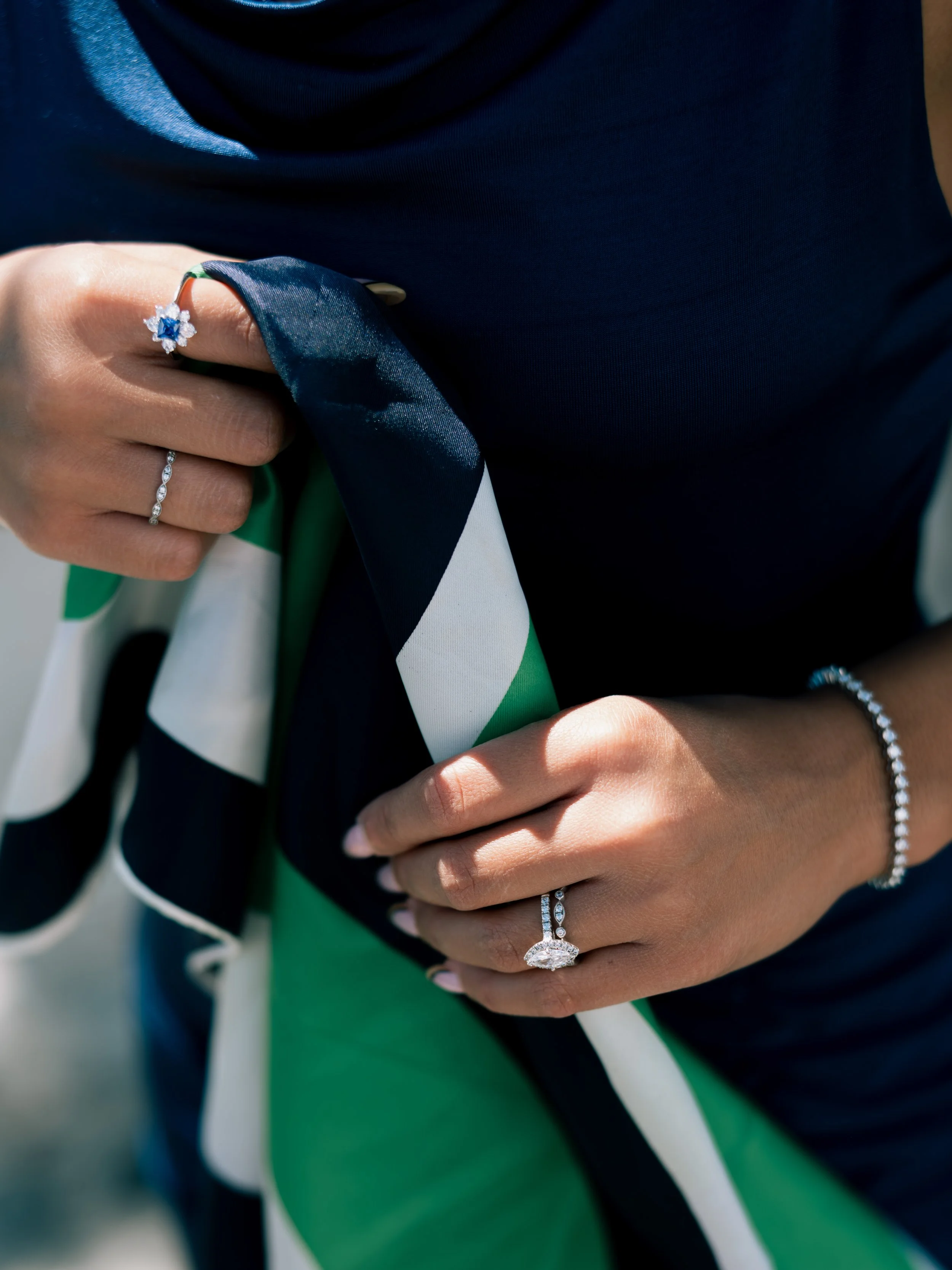 Close-up of a person wearing multiple diamond rings, a diamond bracelet, and a dark-colored top, holding a colorful scarf with green, white, and navy blue stripes.
