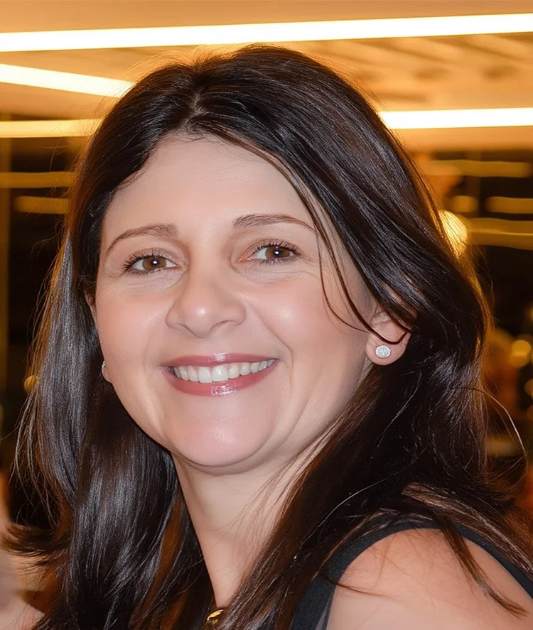Close-up of smiling woman with dark brown hair, light skin, earrings, in an indoor setting with warm lighting