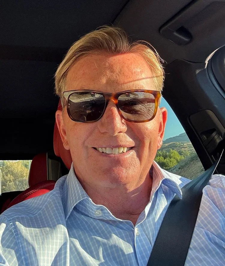 A man with blonde hair wearing sunglasses and a light blue dress shirt, sitting in the driver's seat of a car, smiling.