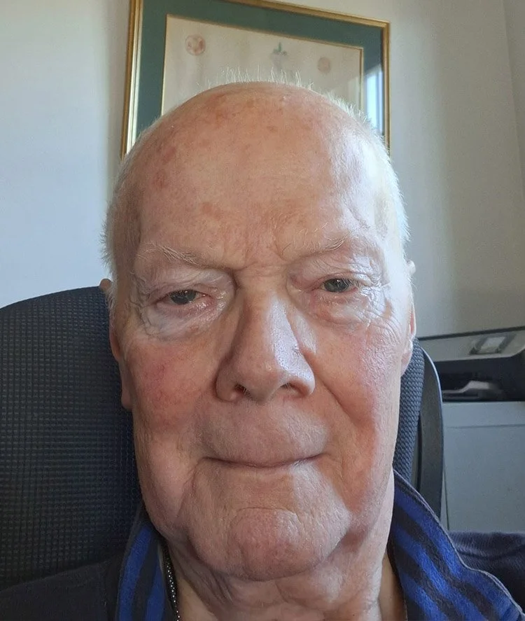 A close-up of an elderly man with light-colored skin, bald head, and wrinkled face, sitting indoors with a framed picture in the background.