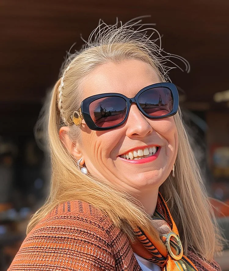 A woman smiling, wearing large sunglasses, a patterned blazer, and a colorful scarf, with a blurred outdoor background.