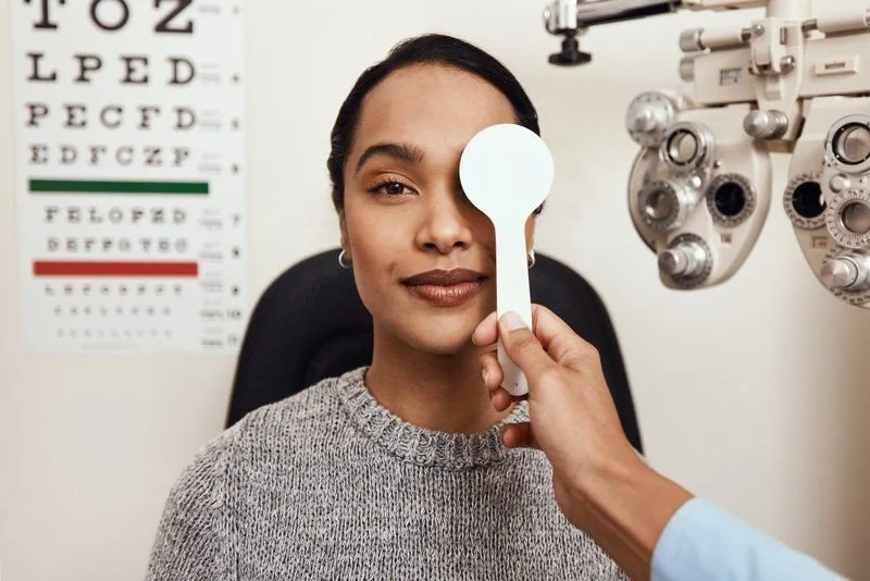 Modella durante un esame oculistico in uno studio optometrico, con una tabella otoscopica sullo sfondo e strumenti professionali di optometria.