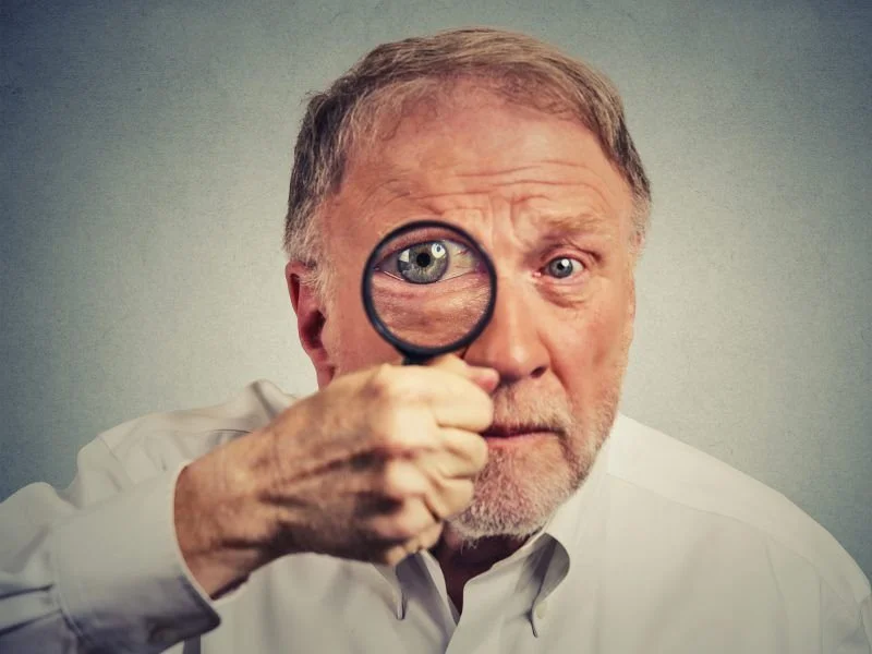 Uomo con capelli grigi, barba, vestito bianco, che tiene una lente di ingrandimento davanti a un occhio