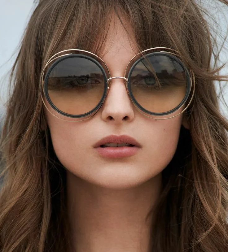 Ragazza con acconciatura riccia e occhiali da sole rotondi di grandi dimensioni, sfondo naturale.