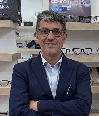 Uomo sorridente con occhiali, vestito con giacca blu e camicia bianca, in negozio di occhiali con scaffali di occhiali sullo sfondo.