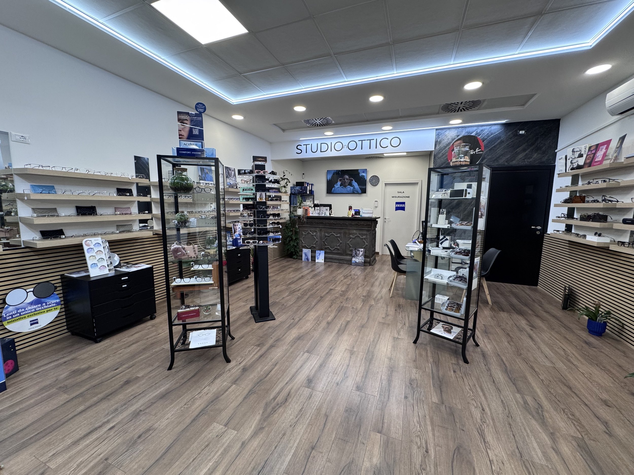 Interno di un negozio di occhiali chiamato Studio Ottico, con esposizione di occhiali su scaffali e vetri espositivi, reception con scrivania e sedie, e dettagli di arredo moderno.