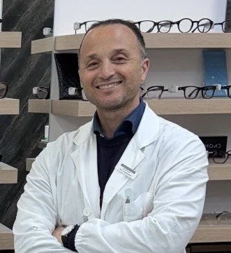 Uomo con camice bianco che sorride in negozio di occhiali, scaffali con occhiali da vista sullo sfondo.