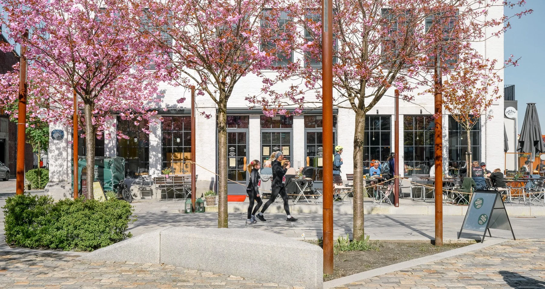 People walking and sitting outside a café with pink flowering trees.