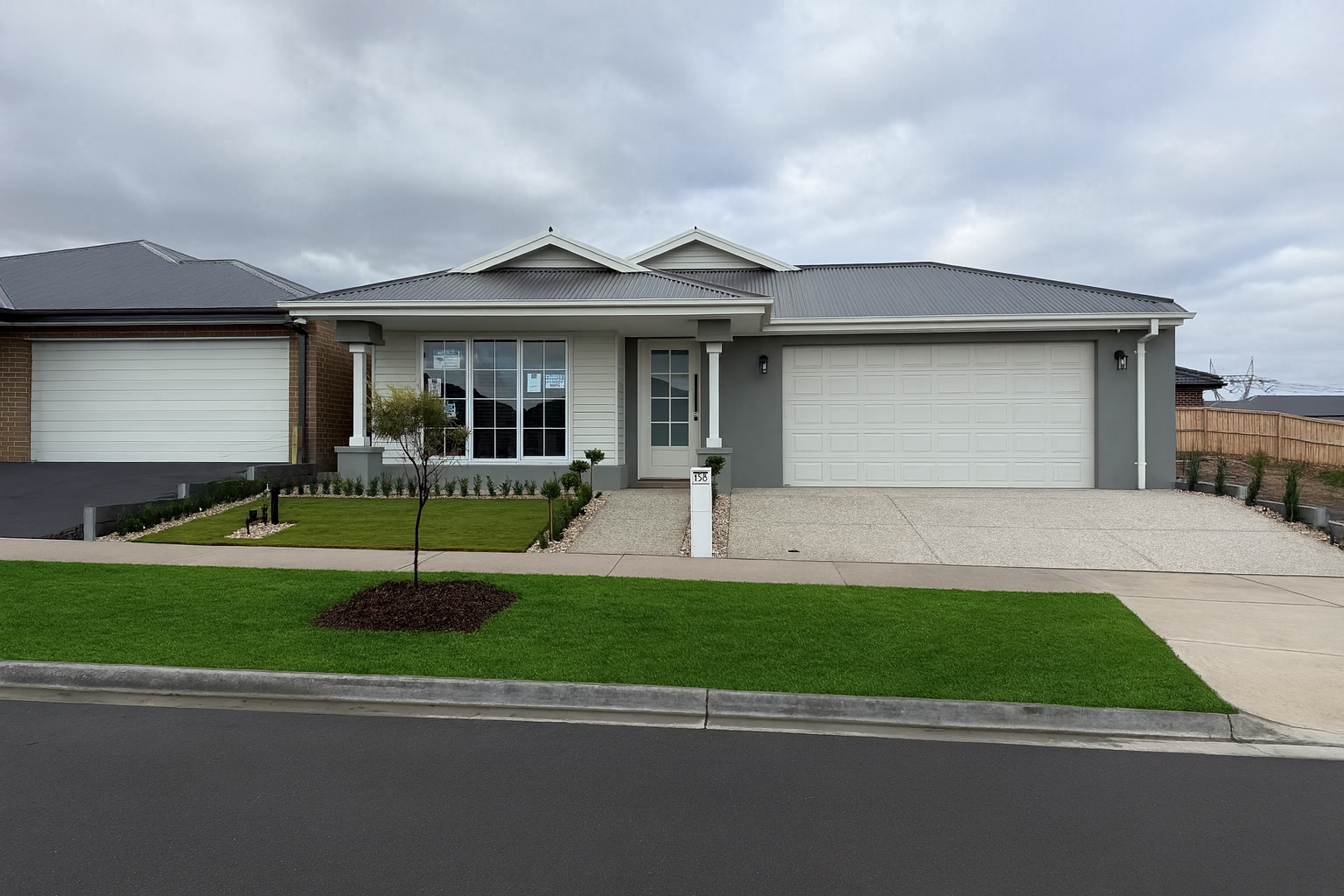 Front view of a modern house with a gray roof, white exterior walls, a two-car garage, a small front yard with a tree, and a well-maintained lawn under a cloudy sky.