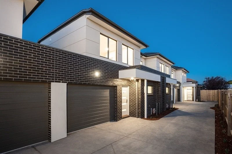 Modern row of townhouses with brick and white siding, illuminated at dusk, featuring attached garages and small front yards.