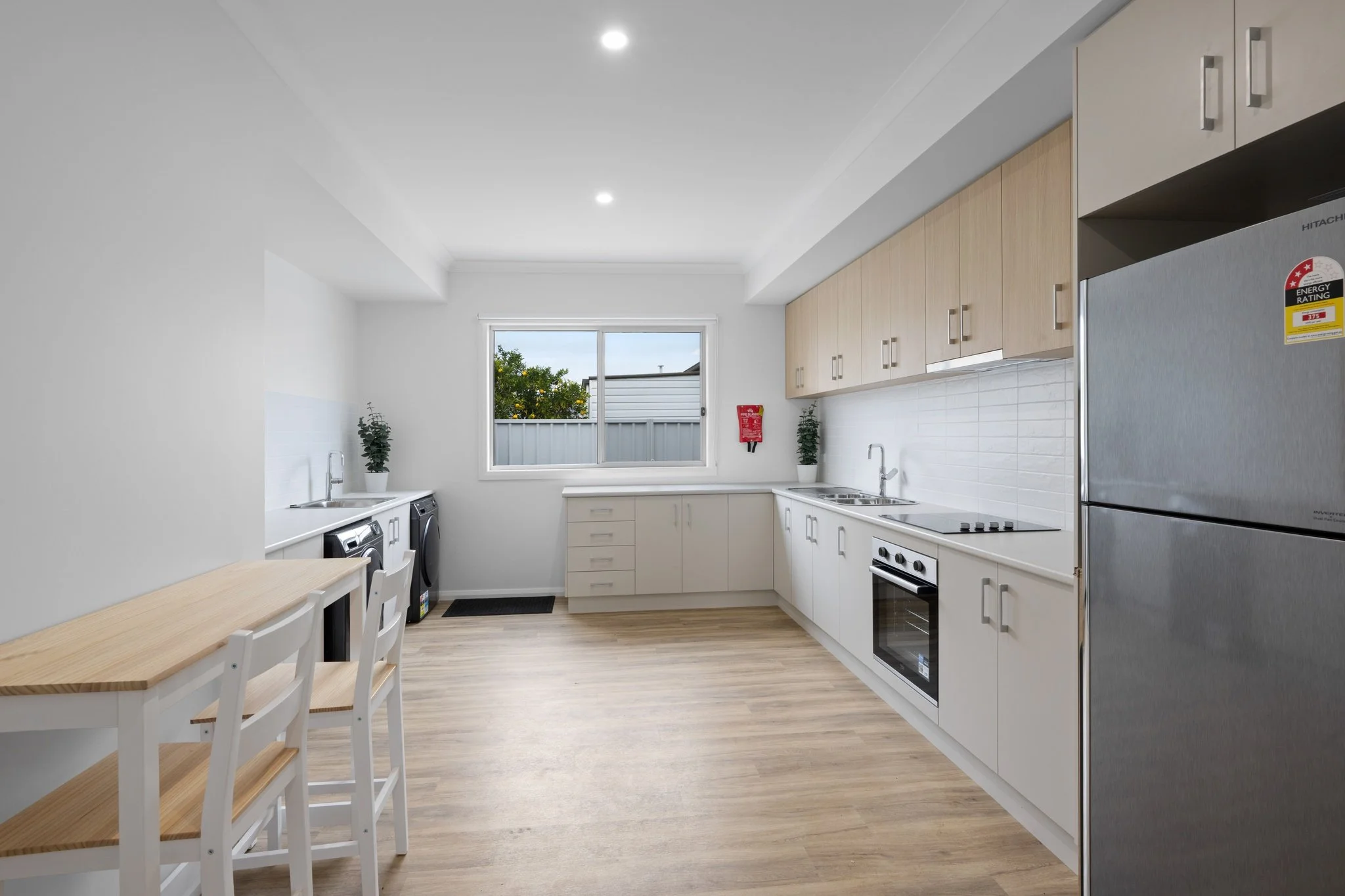 Clean, modern kitchen with white cabinets, a stainless steel fridge, oven, and double sink, wood flooring, a small dining table with two chairs, and a window overlooking greenery.