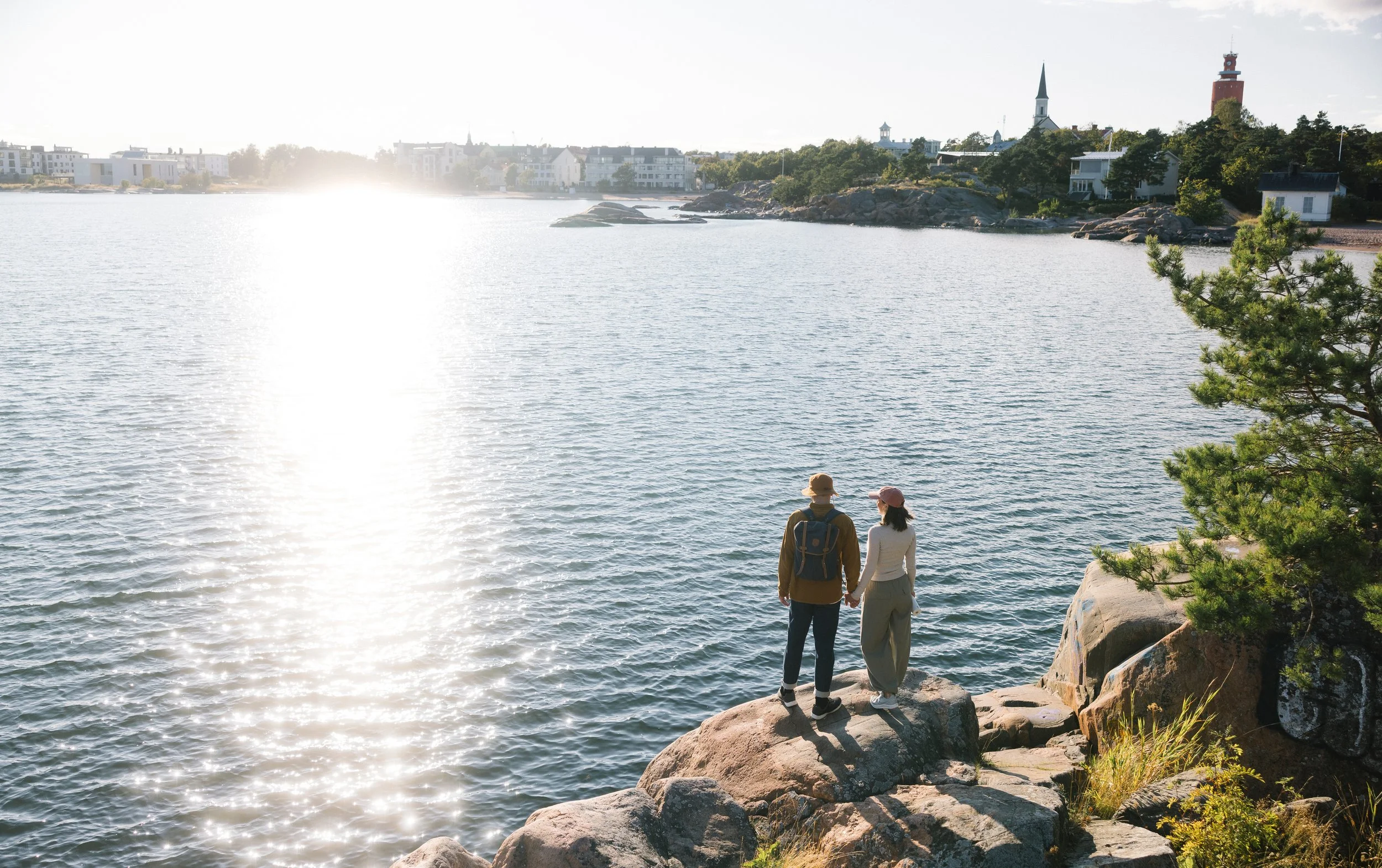 A couple holding hands and standing on rocks by a body of water, with a town and churches in the background, during sunset.